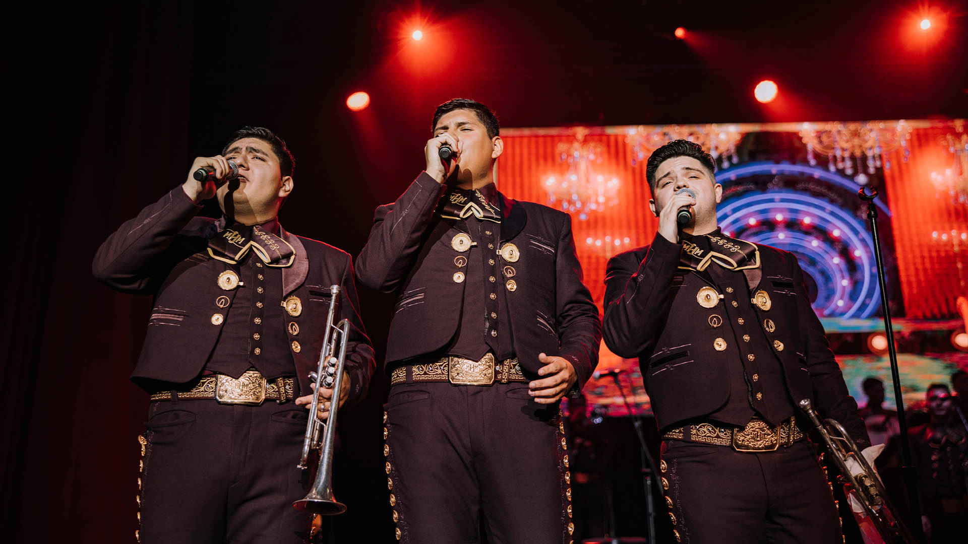 Three Mariachi members in dark outfits. They are hold trumpets in one hand and microphones in the other. All three are singing.