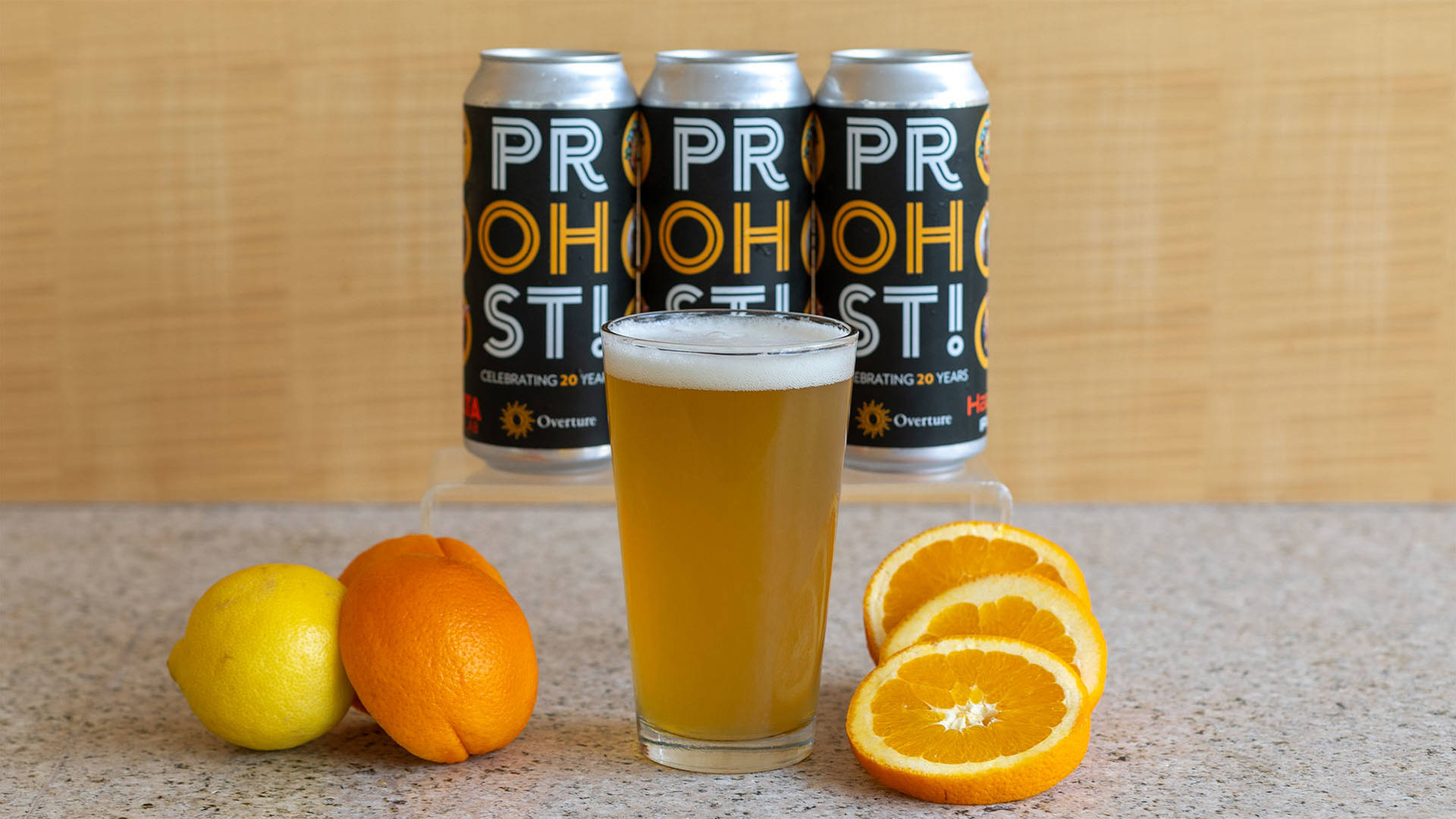 A pint glass of light yellow beer and foam on top on a countertop with sliced orange displayed next to it. Behind are three tall beer cans with black labels that say "PROHST!" in bold yellow and white text.