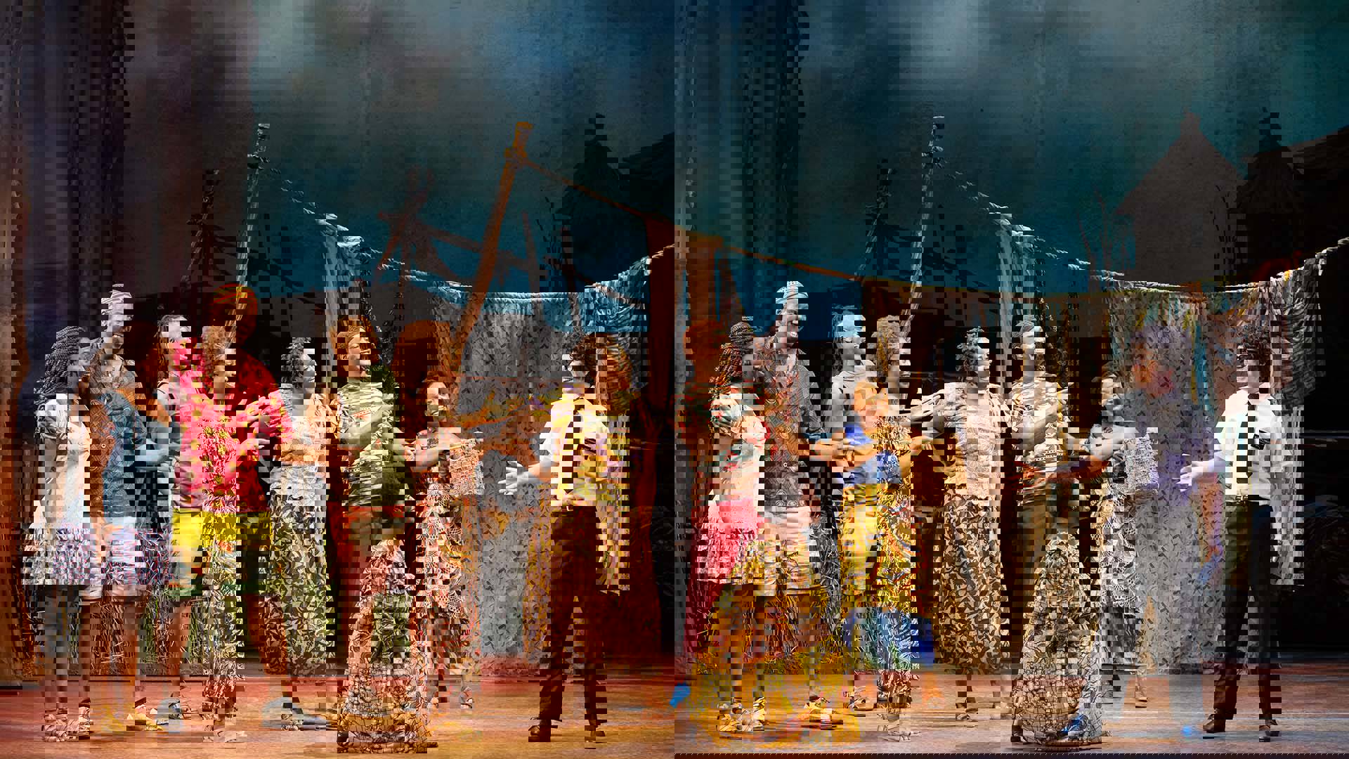 A vibrant ensemble cast- eight people with dark complexion wearing colorful African patterned costumes- interacts on stage with a light complexion man with dark curly short hair and glasses, wearing black pants, a white short-sleeve button up shirt and black tie holding a leather book. Each person holds a dynamic expression bringing the scene to life against a lively village backdrop. 