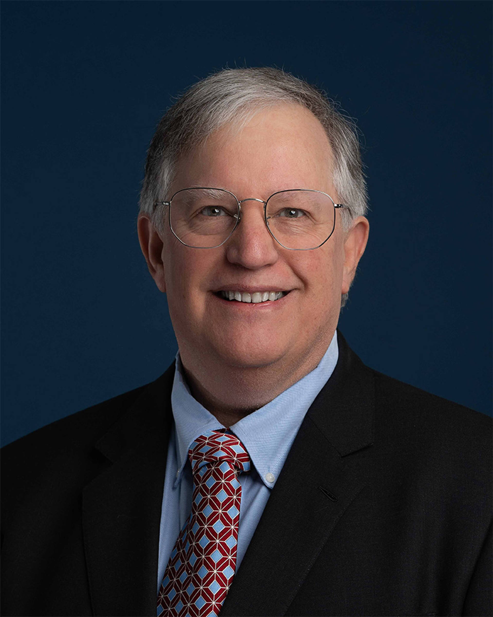 A middle aged white man with grey eyes and grey hair with glasses and a suit.