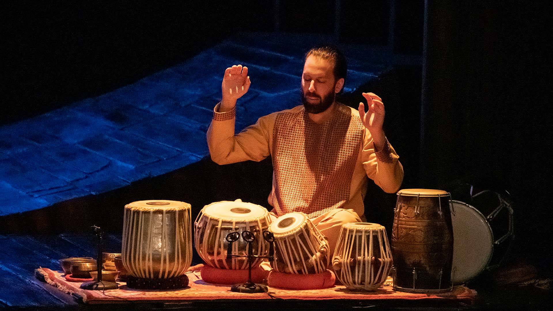 A man with medium complexion, medium dark hair and a dark beard wearing a tan kurta. He is sitting on a carpet playing Tabla, multiple hand drums, on stage.