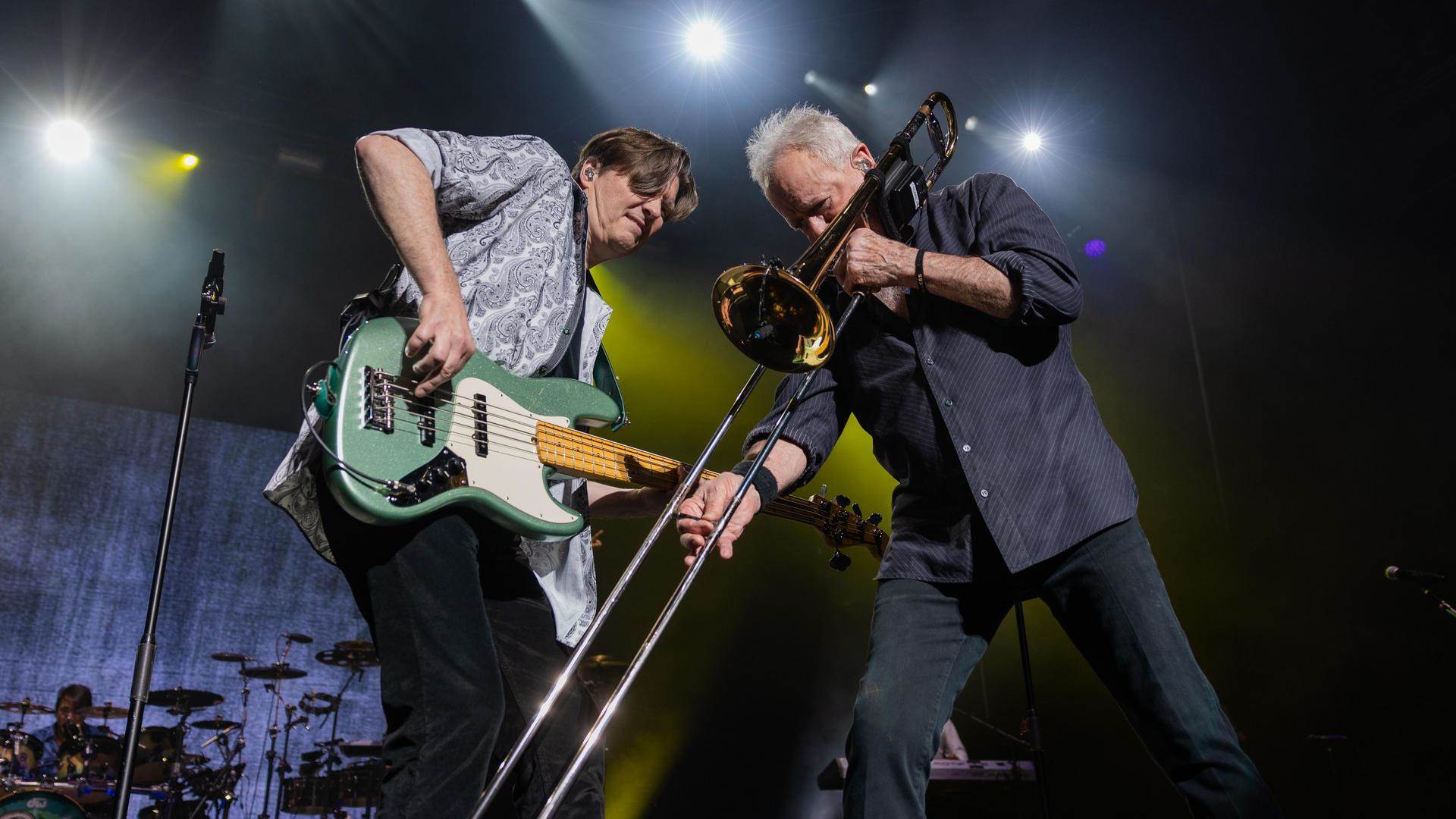 Two older white men on stage. The man on the left is playing a green guitar and the man on the right is playing a trombone.