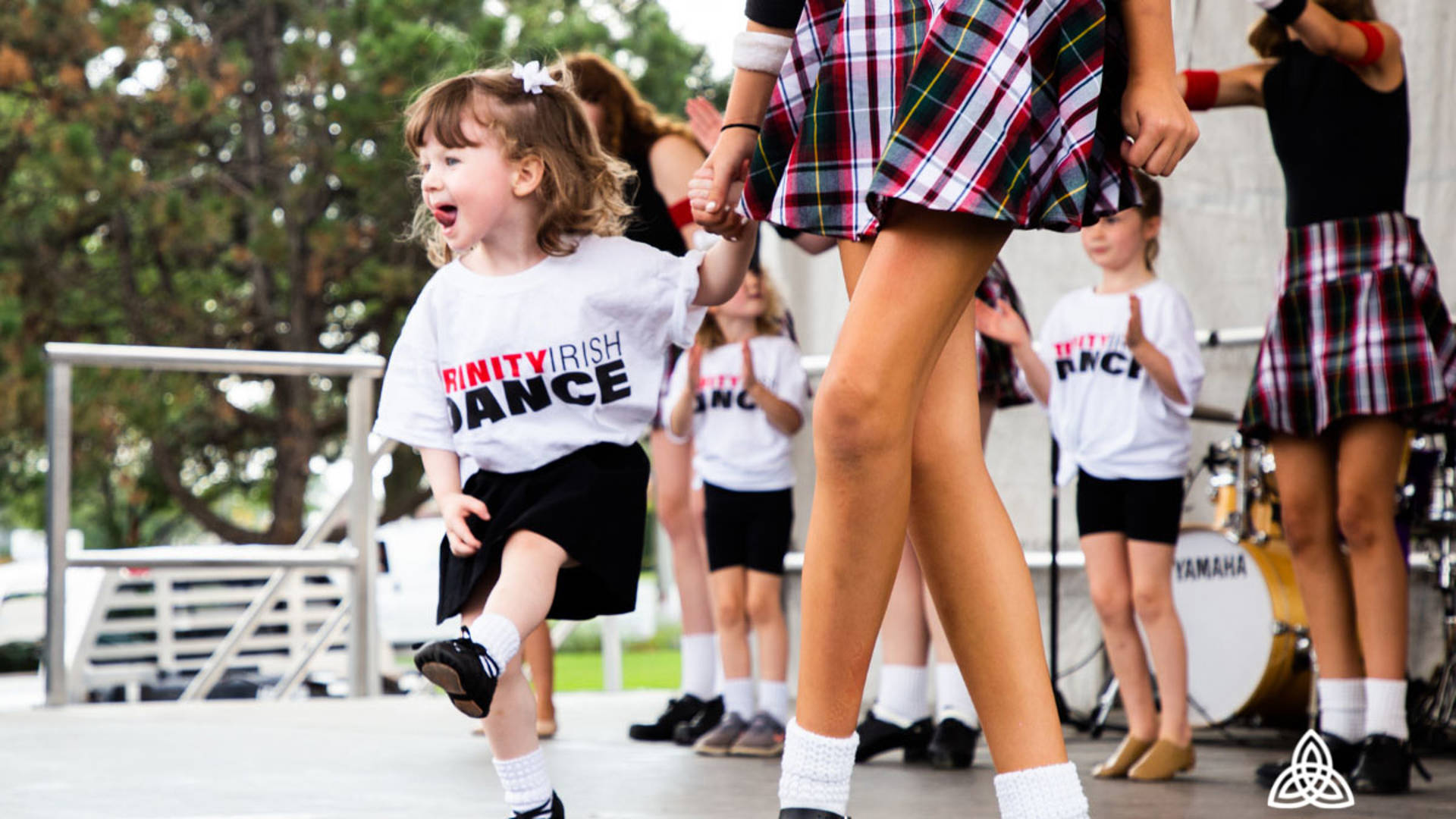 Trinity dancers performing on stage. Focus on a young girl with brown hair wearing a white t-shirt and black skirt.