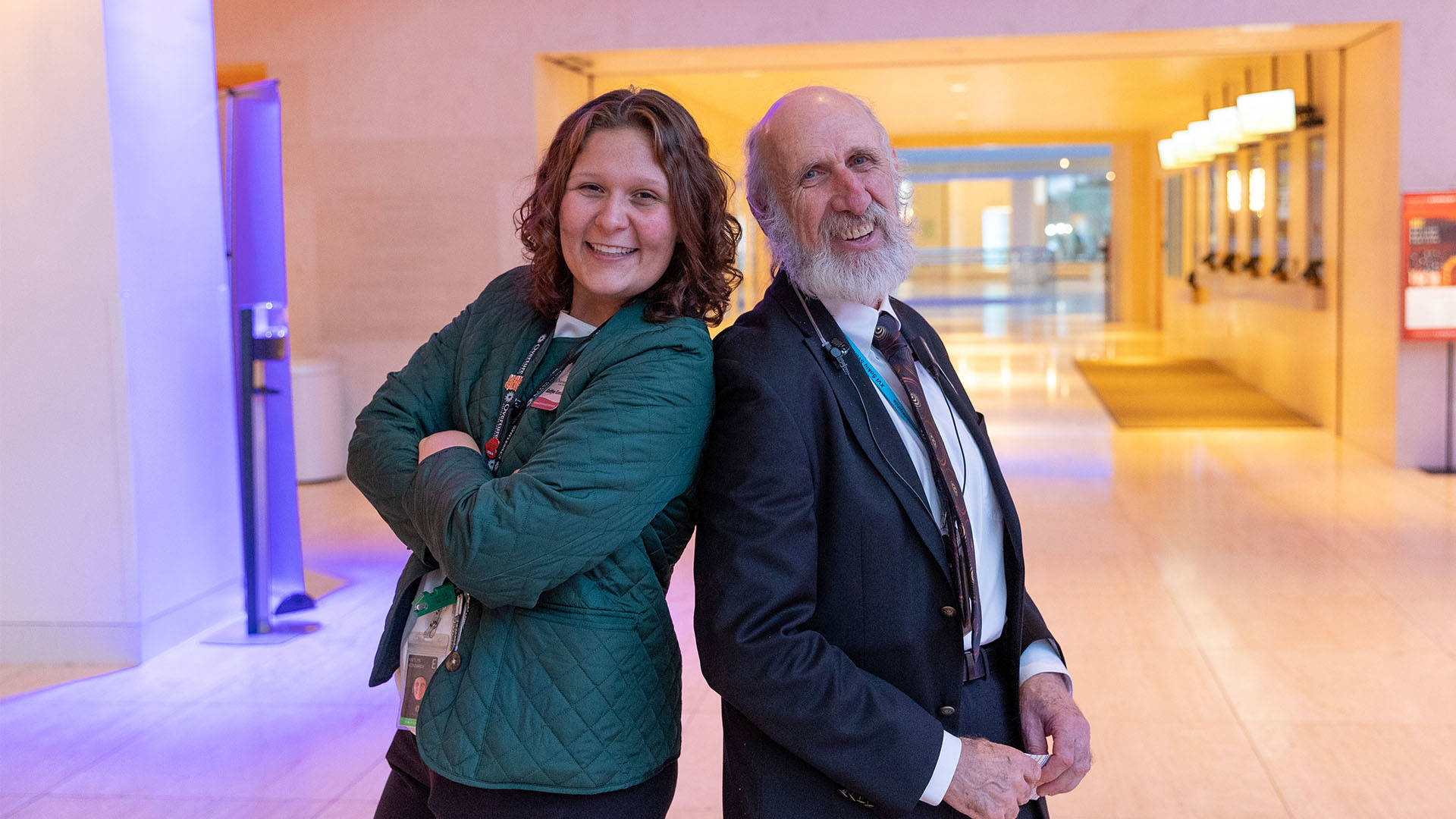 a young caucasian woman in a green jacket stands back to back with an older man with a grey beard. They are in a beige lobby.