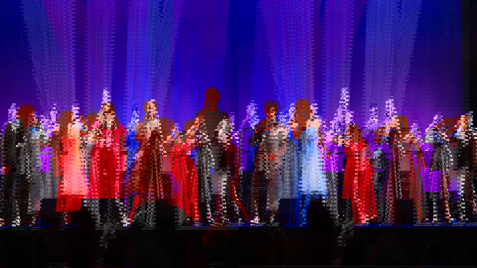 The Jerry Ensemble performing on stage. A group of a dozen teenagers in formal outfits on stage with microphones and a blue backdrop.