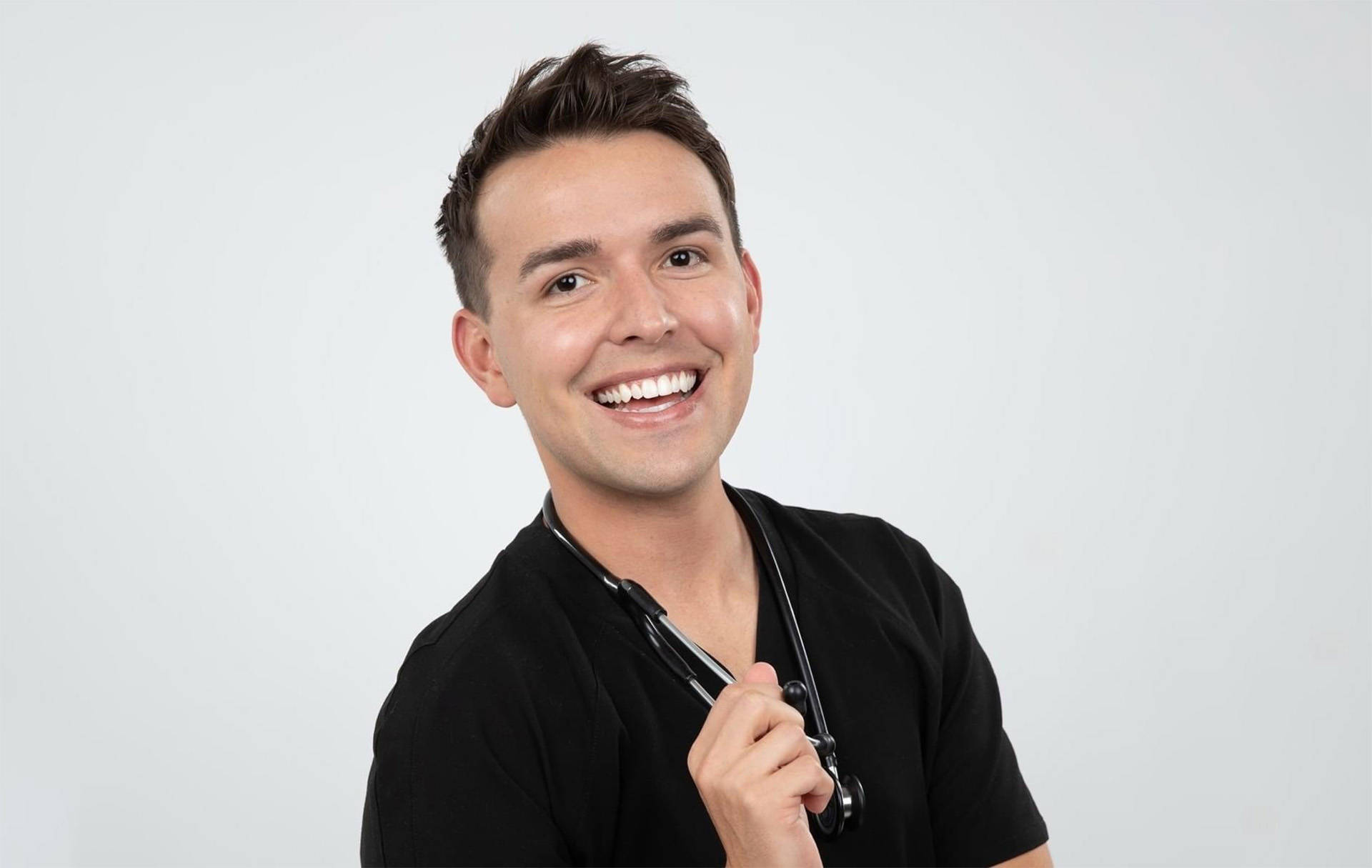 A caucasian man with short black hair smiling. He is wearing a black scrubs top with a stethoscope hanging from his neck.