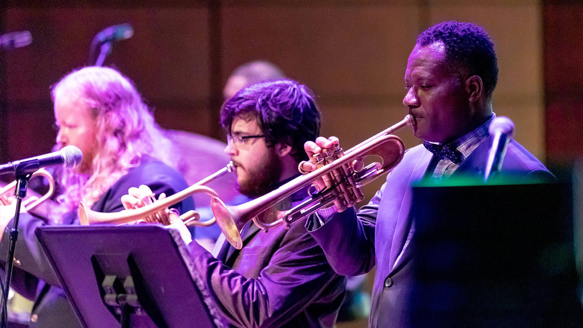 Three people sitting in front of music stands playing trumpets. They are wearing blue suits.