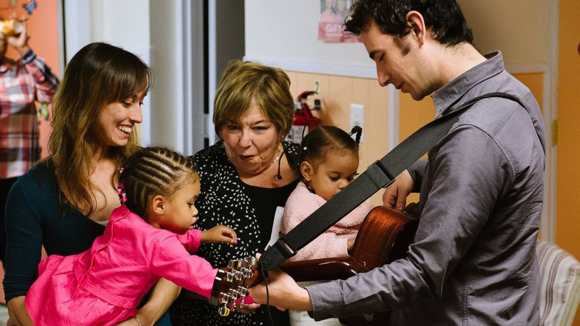 A man playing the guitar to two young African-American children being held by adults.