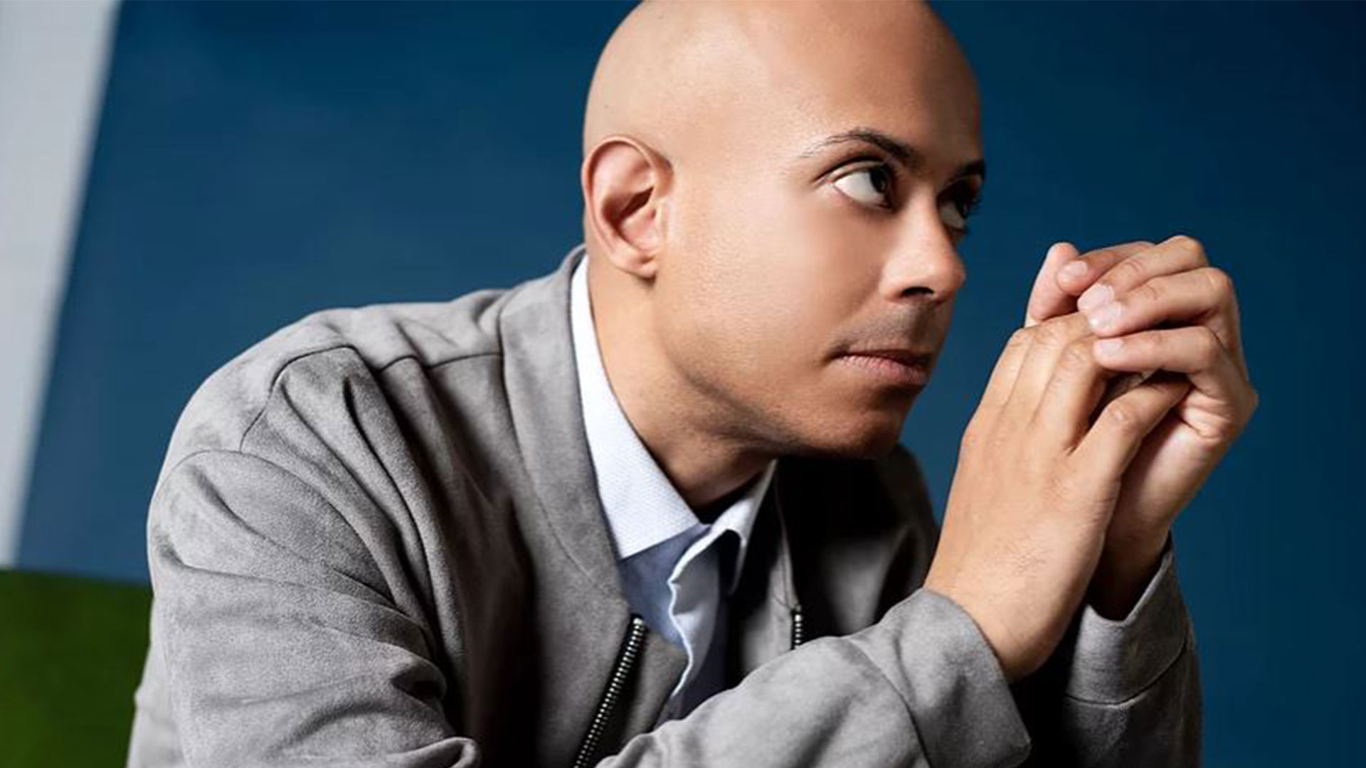 A young african american man with with a bald head is looking sideways and holding his hands together in front of his face. He has a light blue collared shirt on and a grey velvet jacket.