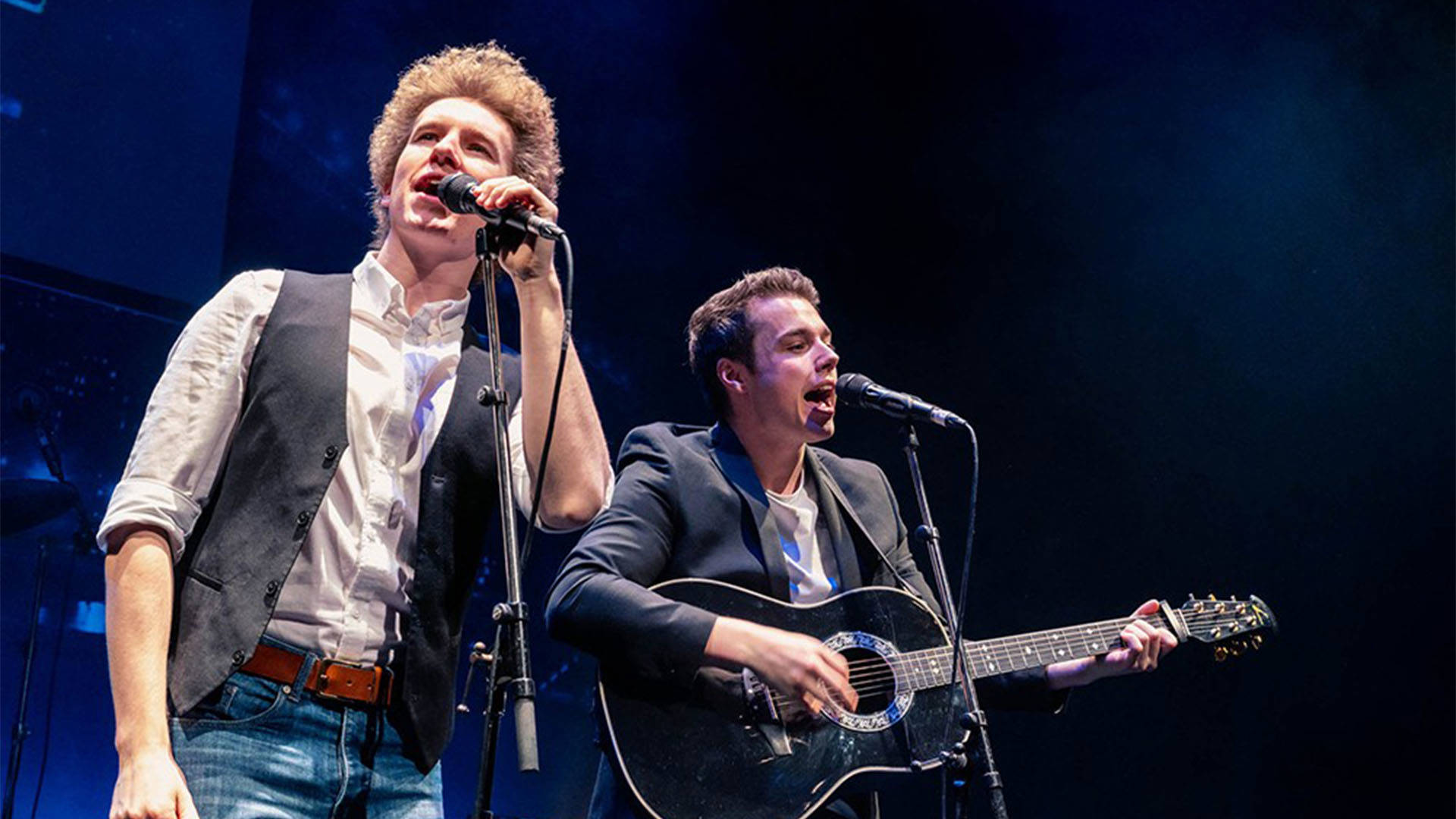 Two white men standing behind microphones. The one on the left has light, curly hair and is wearing a grey vest and white shirt. The one on the right has short, brown hair, is wearing a grey jacket and playing a black guitar.