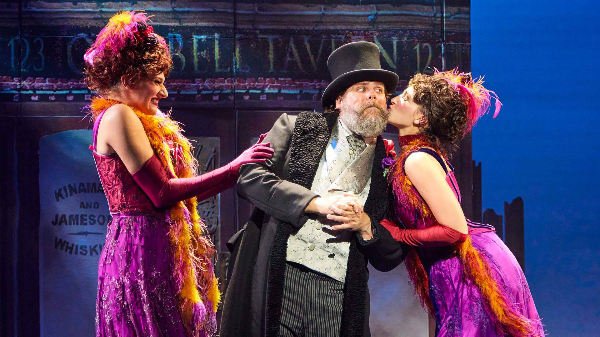 Two women in pink dresses and feather boas stand next to a man with a top hat and gray beard. On woman is kissing the man on the cheek.