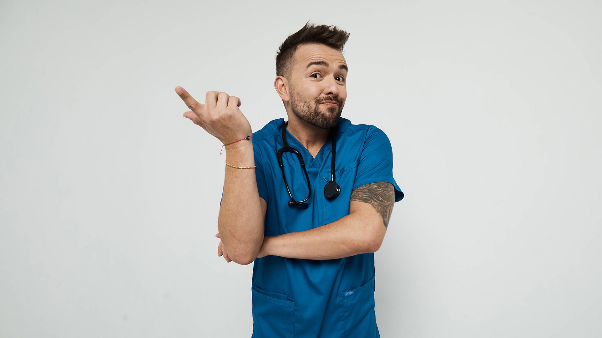 A smiling white man with a beard, short styled brown hair wearing blue scrubs with a stethoscope around his neck. He is smiling mid-shrug.