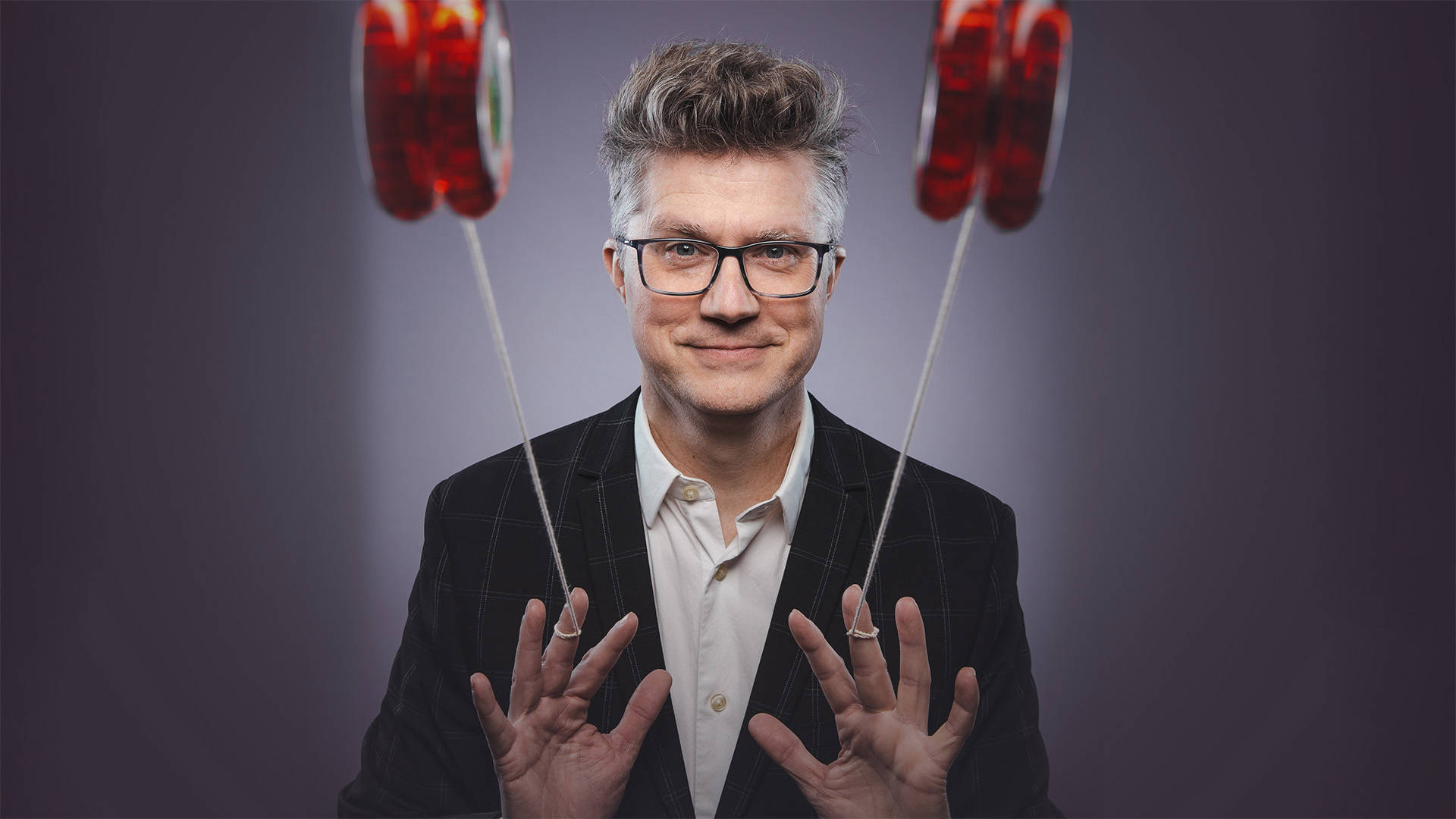 A white man with short tossed hair and glasses in a suit jacket and white dress shirt looks towards the viewer. Both his hands are up and he is flicking red yo-yos towards the viewer.
