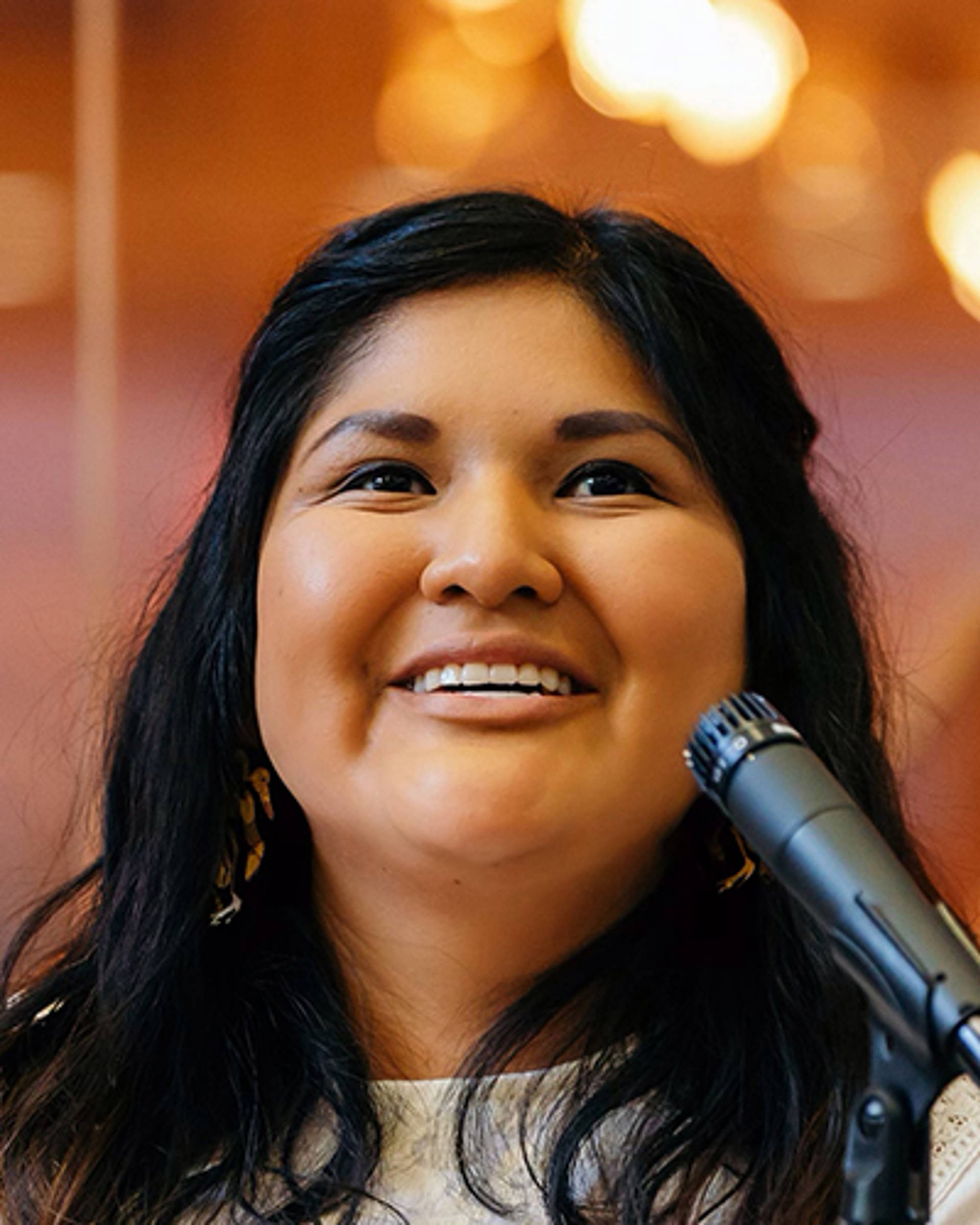 Headshot of a woman of first nations decent with dark hair and dark eyes. She is speaking into a microphone.