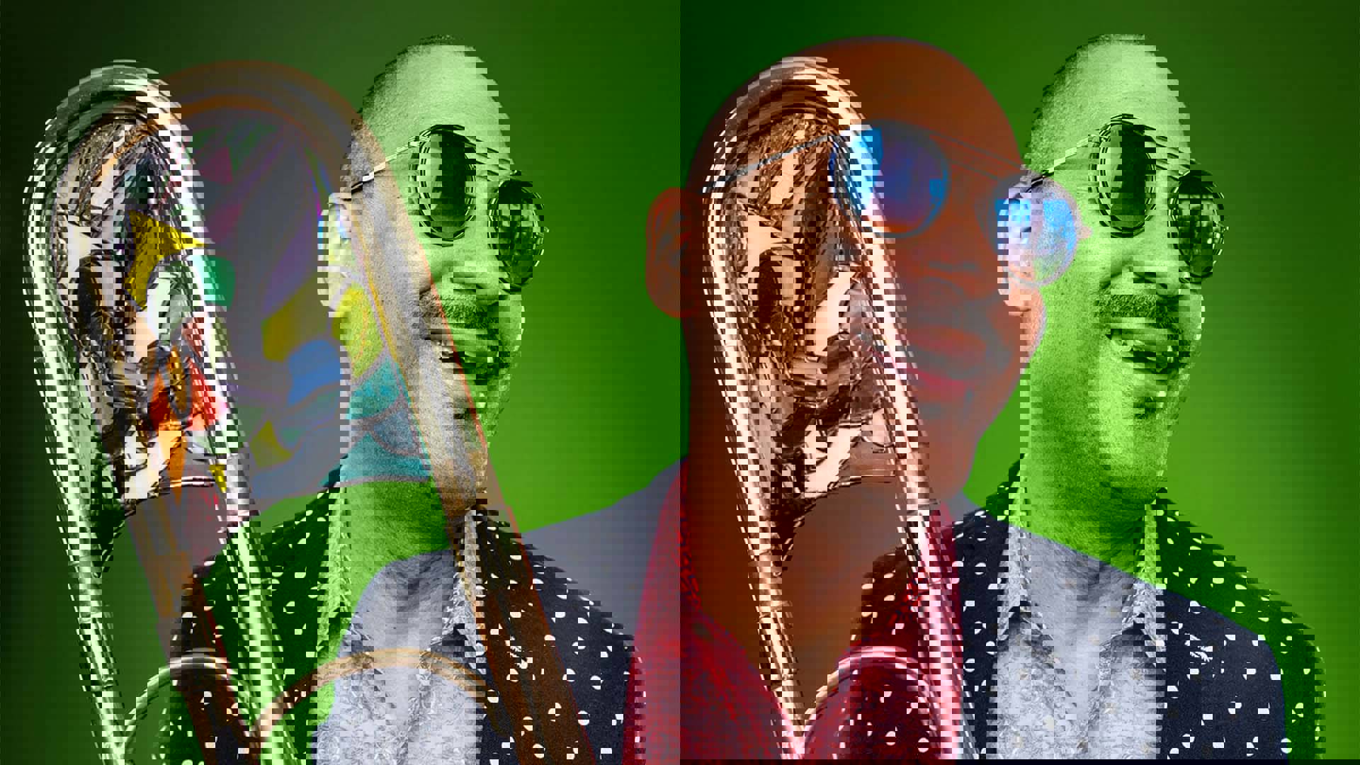 A Black man with buzzed hair, a cropped mustache and sunglasses wearing red dress shirt and polka dotted suit jacket, holding a trombone and smiling.