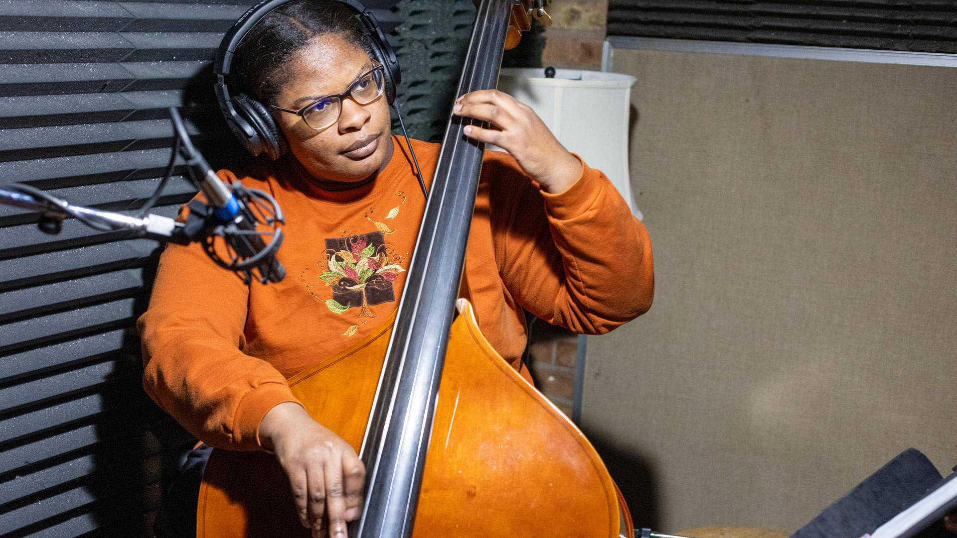A young woman with medium complexion in an orange shirt playing an upright bass. She has headphones on and there is a microphone recording her.