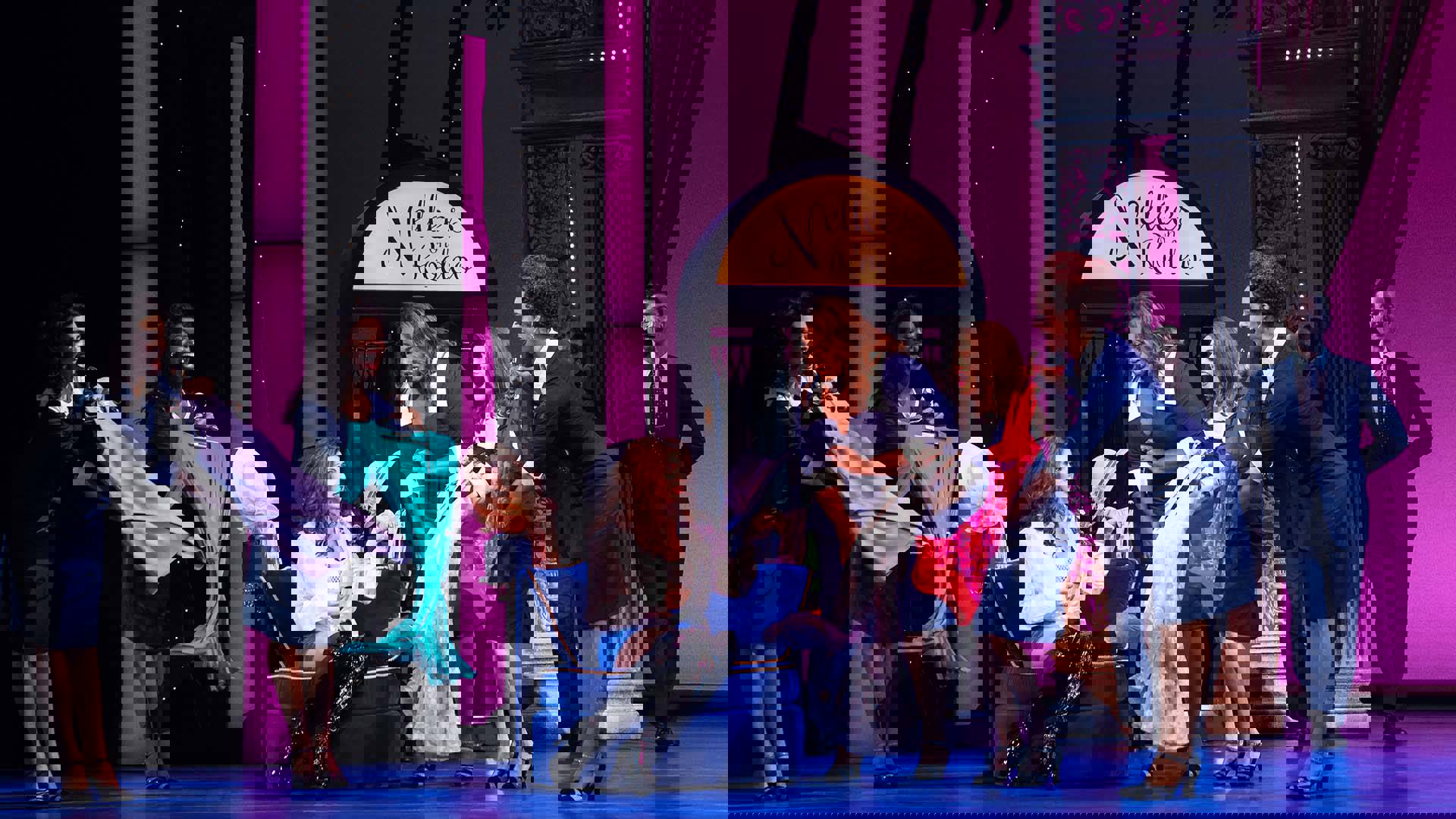 The Cast of Pretty Woman performing on stage. A woman is sitting on a blue, velvet chair. She is surrounded by other women holding fancy dresses.
