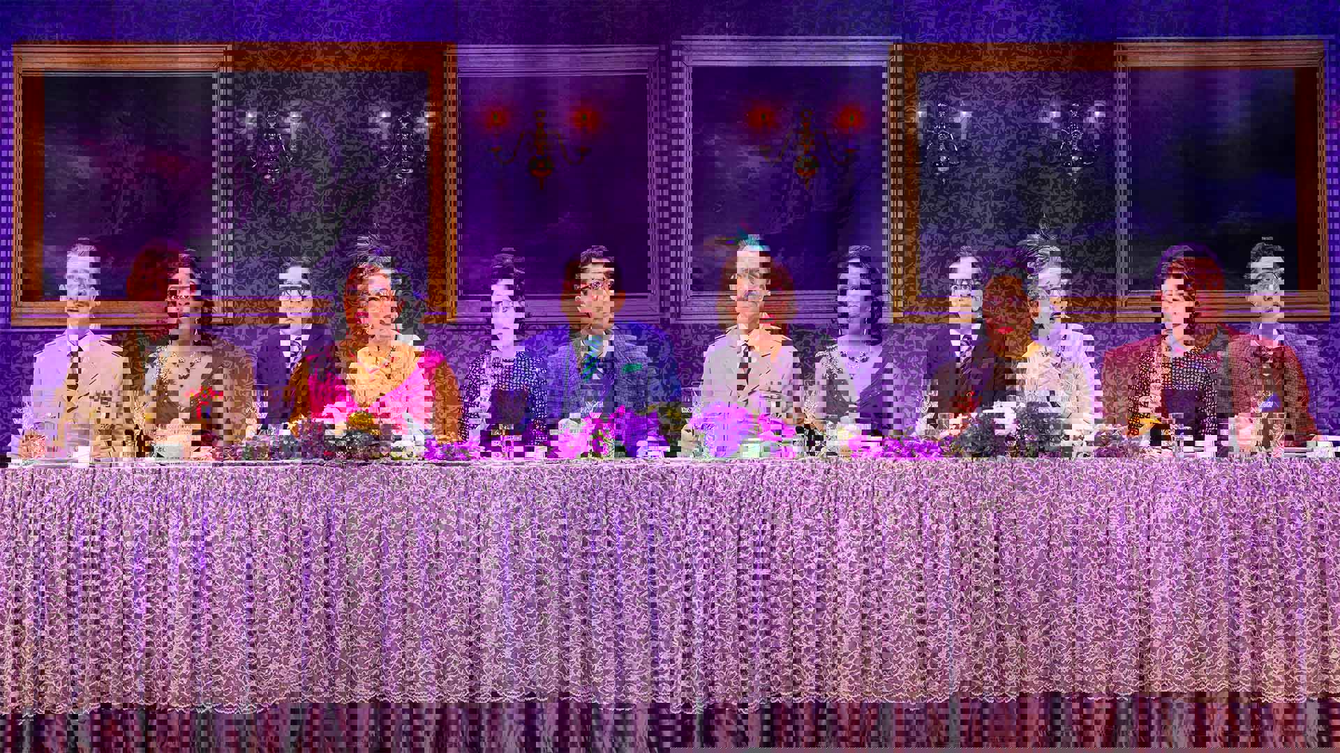 Six actors in formal attire sit all on the same side behind a long fancy dinner table. Each actor is staring forward into the distance with a shocked and perplexed look. 