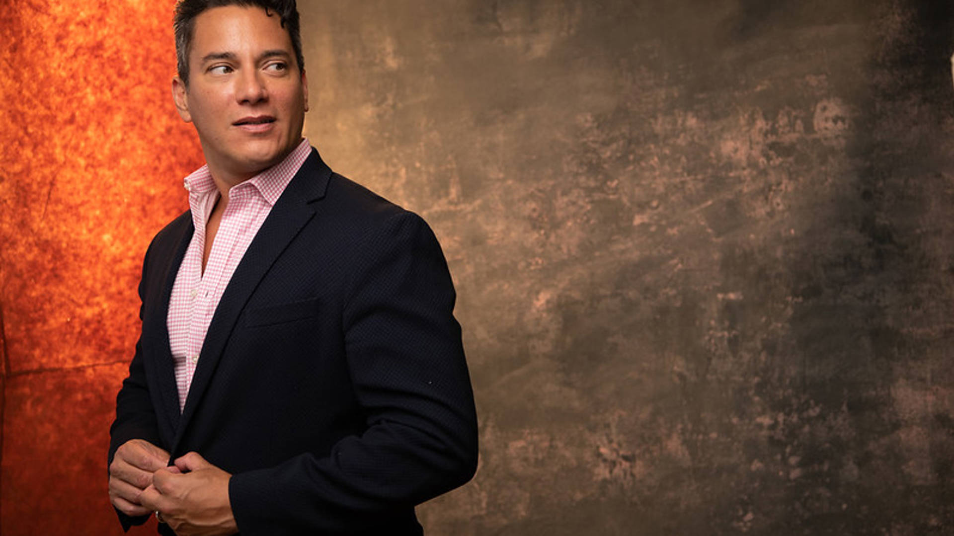 A muscular man with short, dark hair wearing a dark jack and pink dress shirt against a brown and orange background.