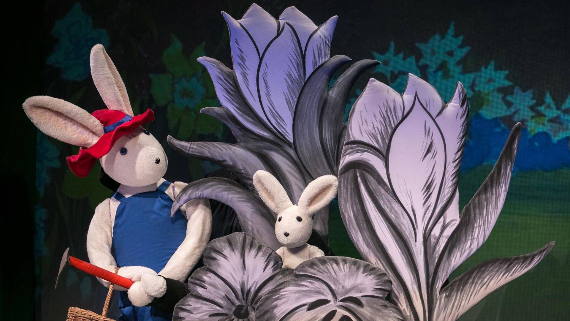 Two white rabbit puppets on stage. One is wearing a red hat and blue apron, the other is much smaller and sitting in a large plant.