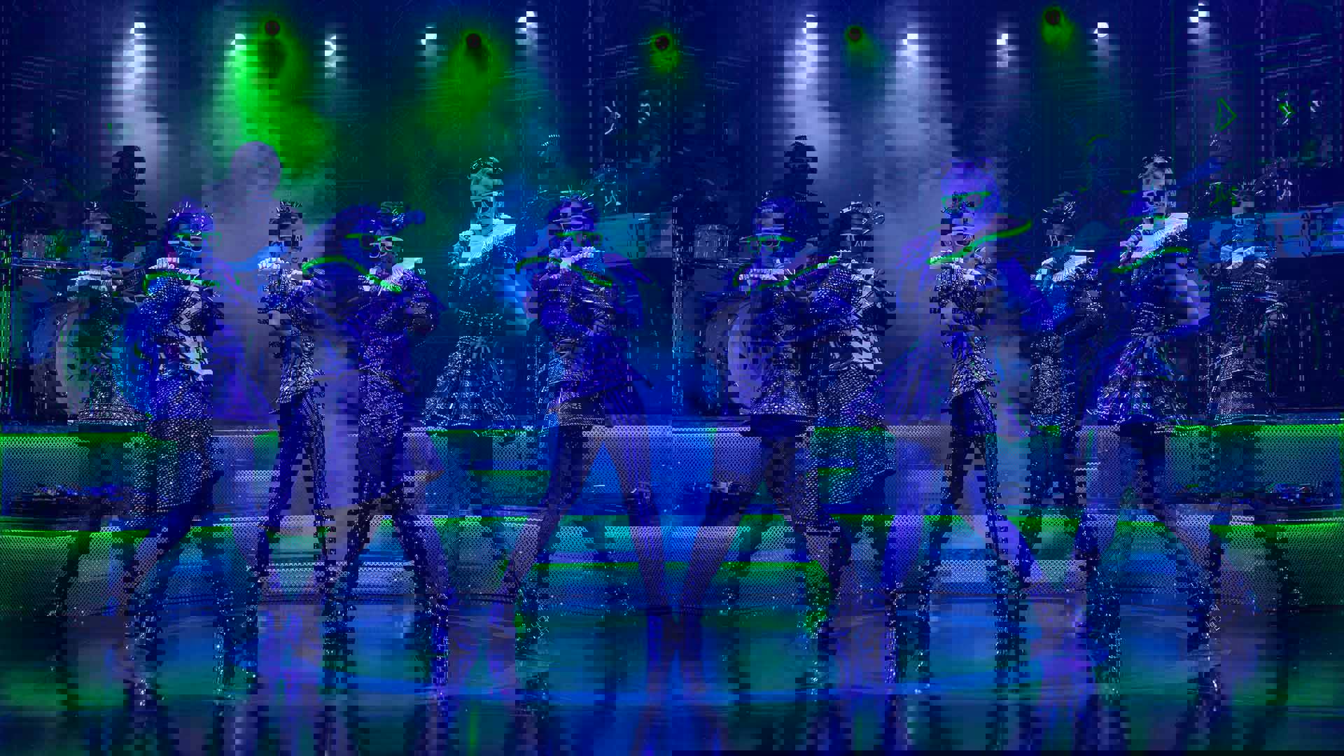 The cast of Six, in sunglasses and futuristic glittery outfits while holding microphones, perform on stage.