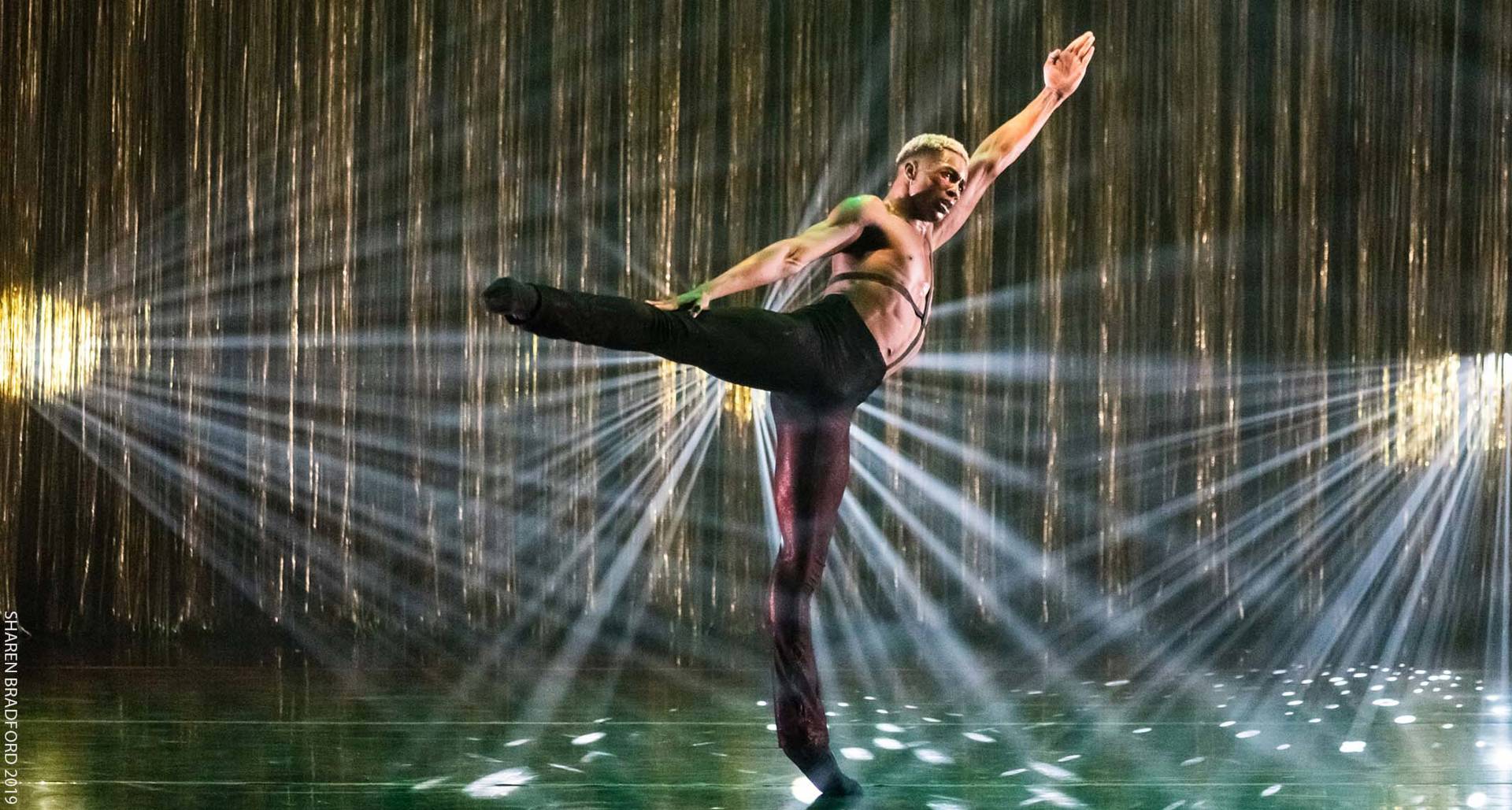 A shirtless Black man with bleached short hair wearing black leggings mid-dance arabesque pose.