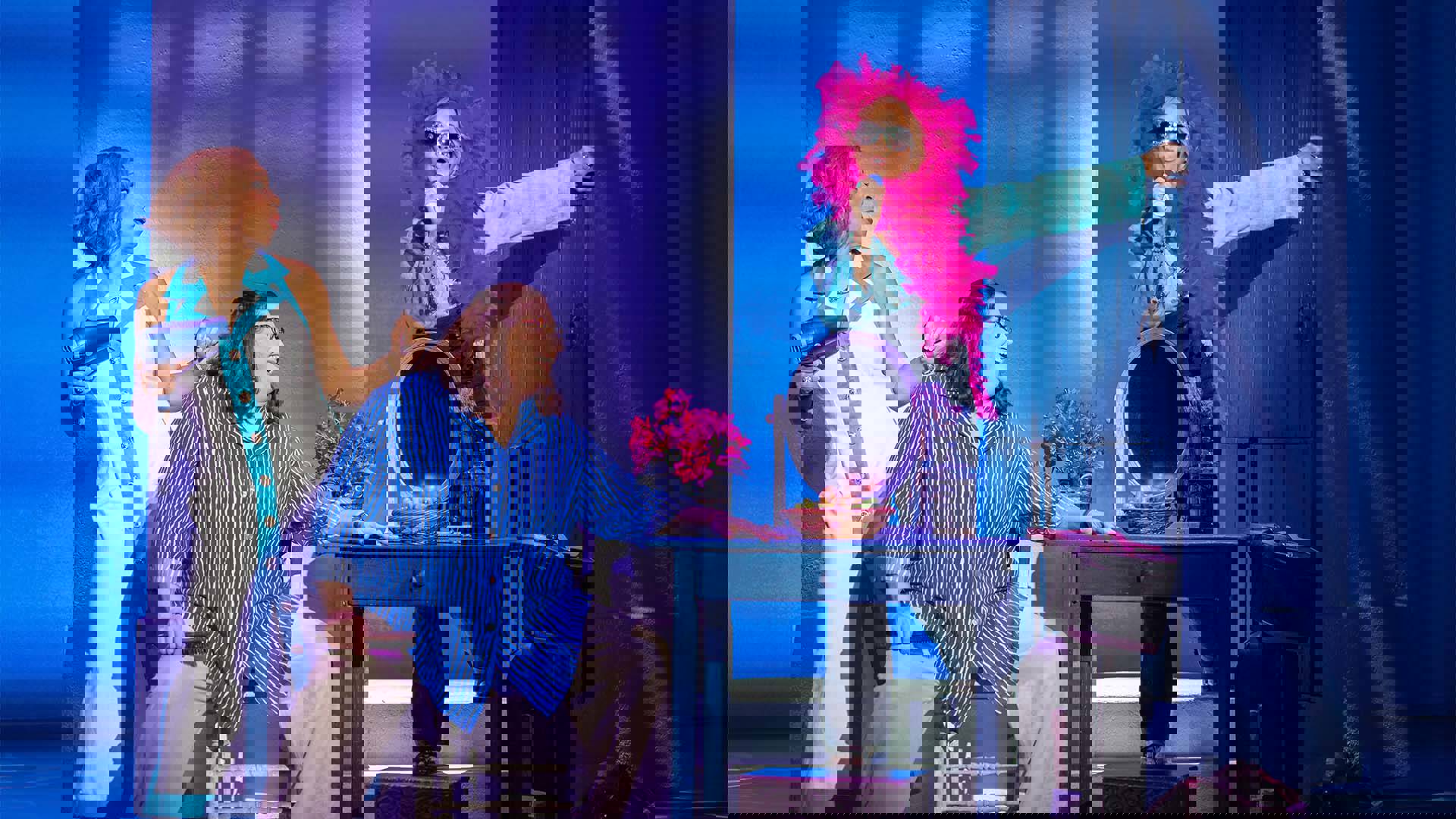 Three women in colorful clothing next to a table and makeup mirror. The woman on the right has a colorful boa around her head.