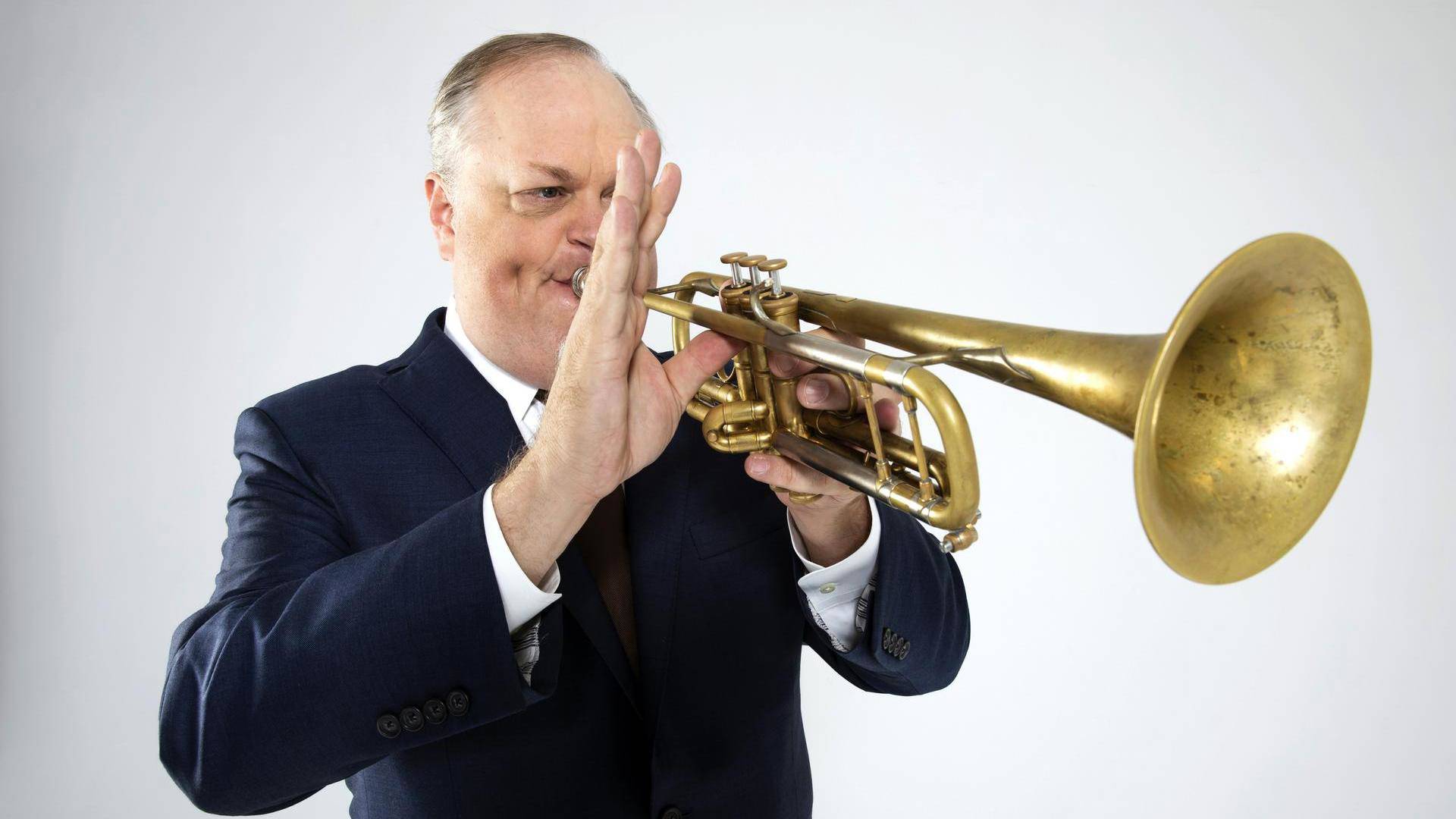 A man with light complexion and grey hair playing a trumpet. He is wearing a black suit against a white background.