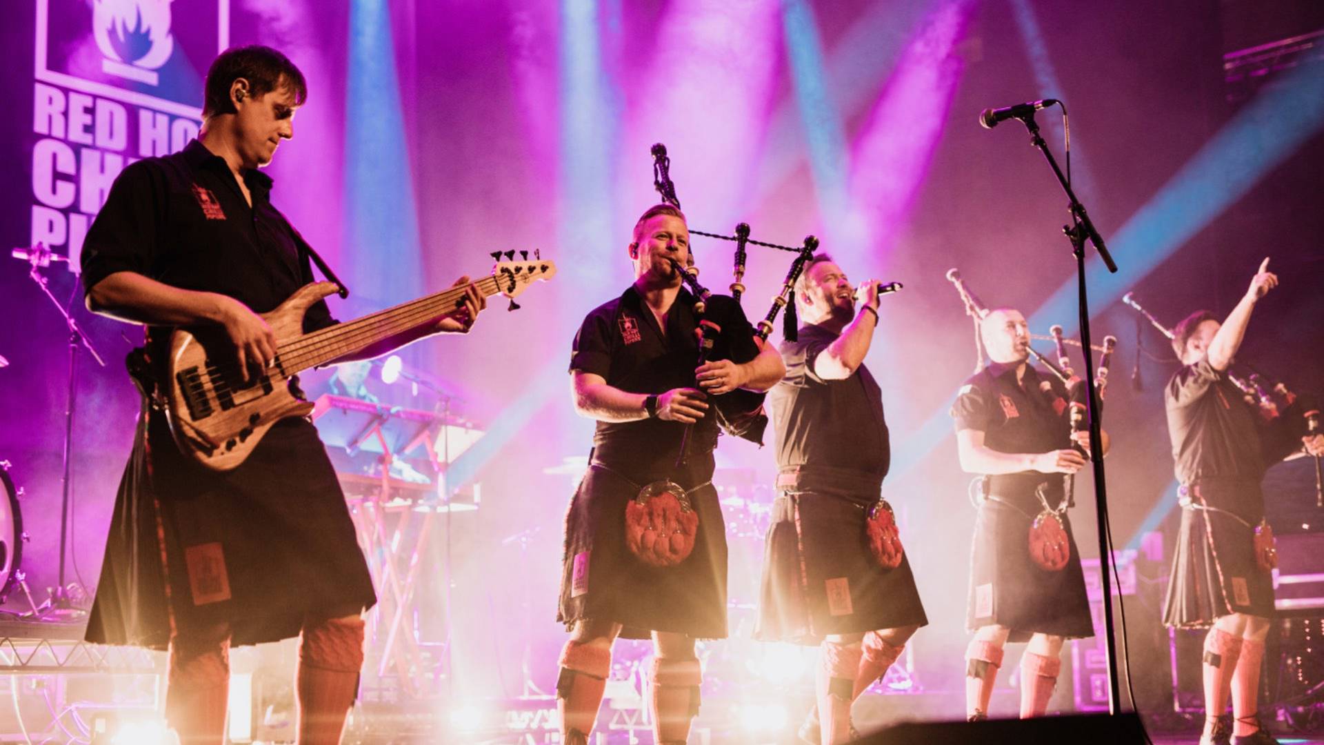 Band members in kilts performing on stage with a bagpiper in the spotlight.