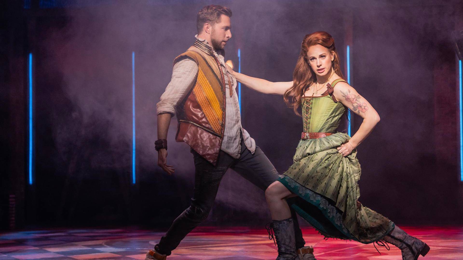 A woman in a green dress with light complexion and reddish hair steps dramatically towards a man with her right hand on his chest. The man, in a brown and yellow vest with light complexion and brown hair, is stepping backward.