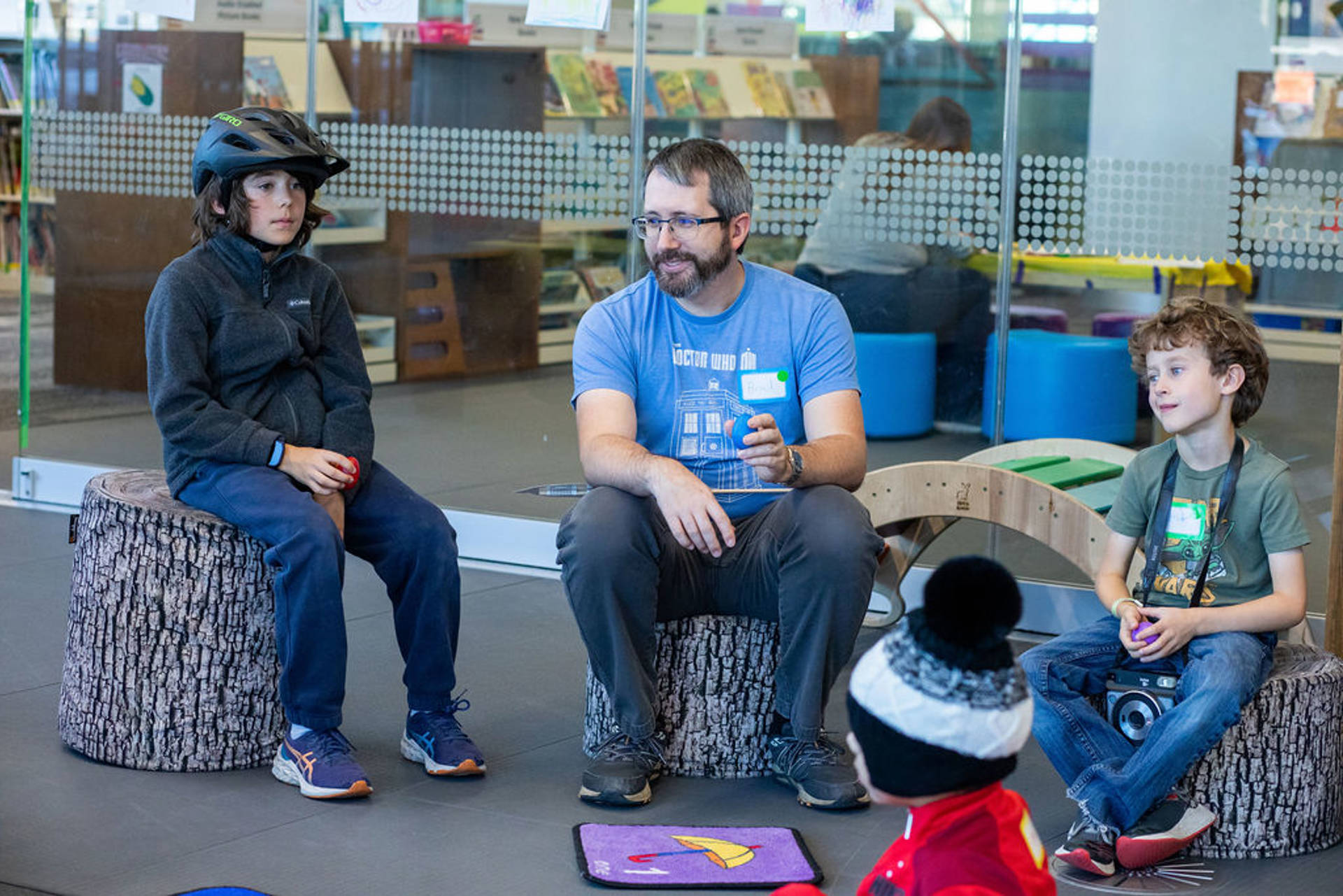 A man with light complexion, greyish hair and glasses sitting and speaking to a few children.