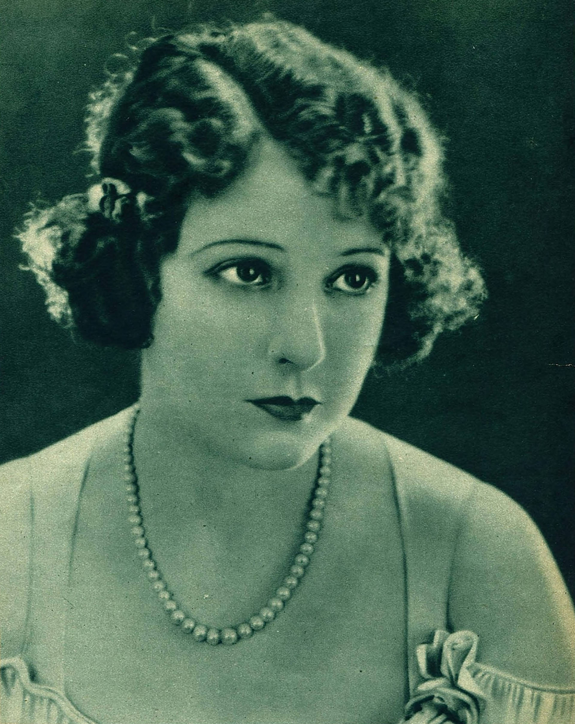 Headshot of a woman from the early 20th century with curly medium length hair, light complexion, a pearl necklace and a dress.