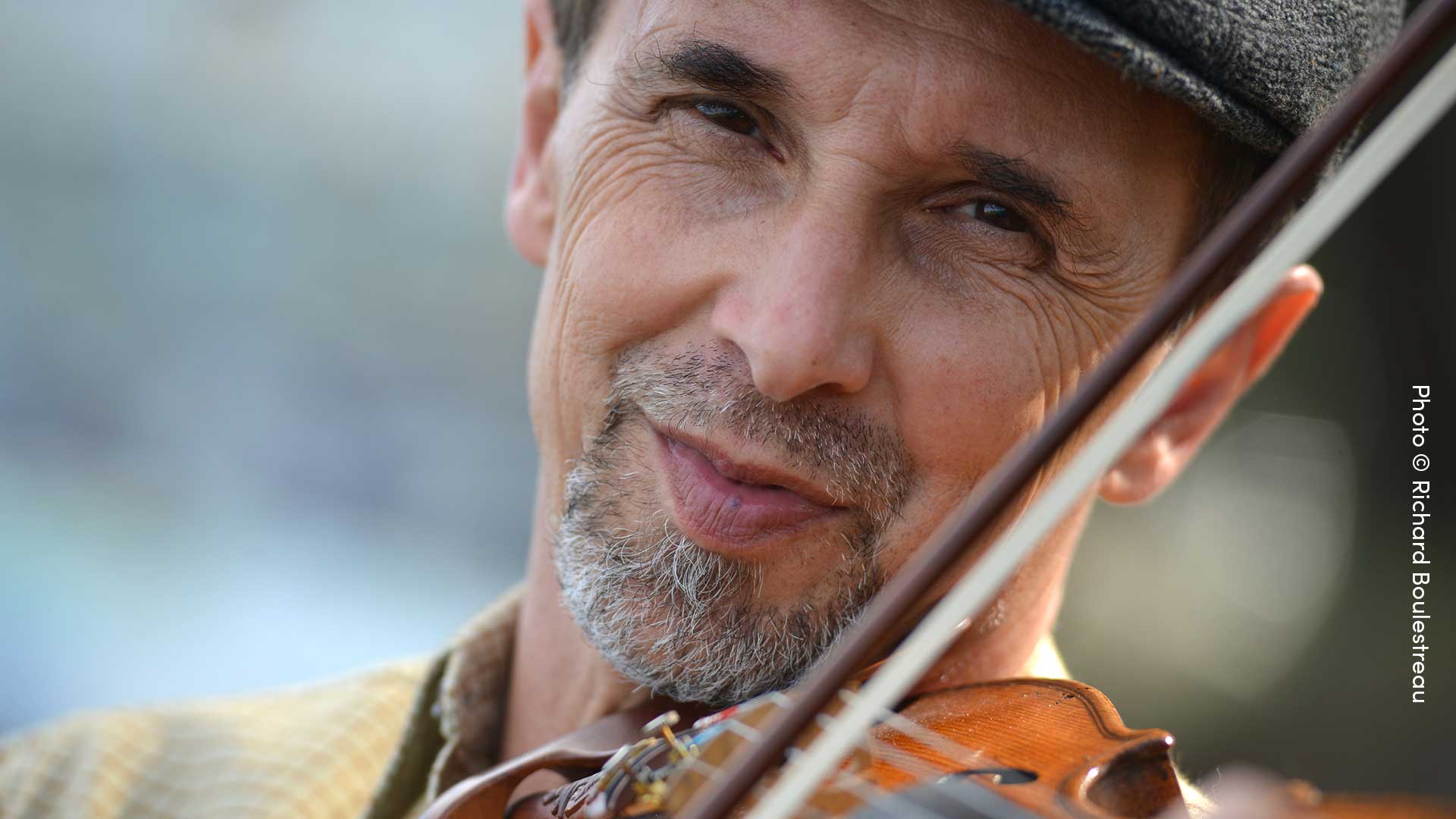 Headshot of a middleaged caucasian man holding a violin. He e has dark eyes and a grey goatee. He is wearing a cap.