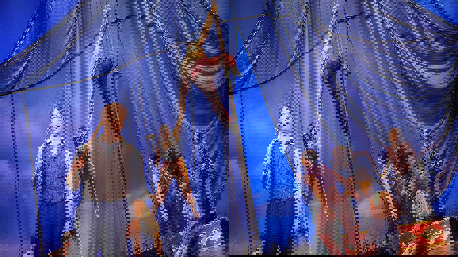 A man with short hair and a light complexion sings while an acrobat holding a sledgehammer swings from above.