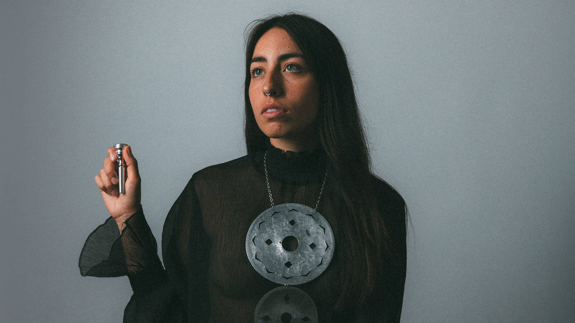 Headshot of a woman with long dark hair, blue eyes and medium complexion in a dark top with a necklace decorated with large metalic discs. She is holding up a trumpet mouthpiece in her right hand.