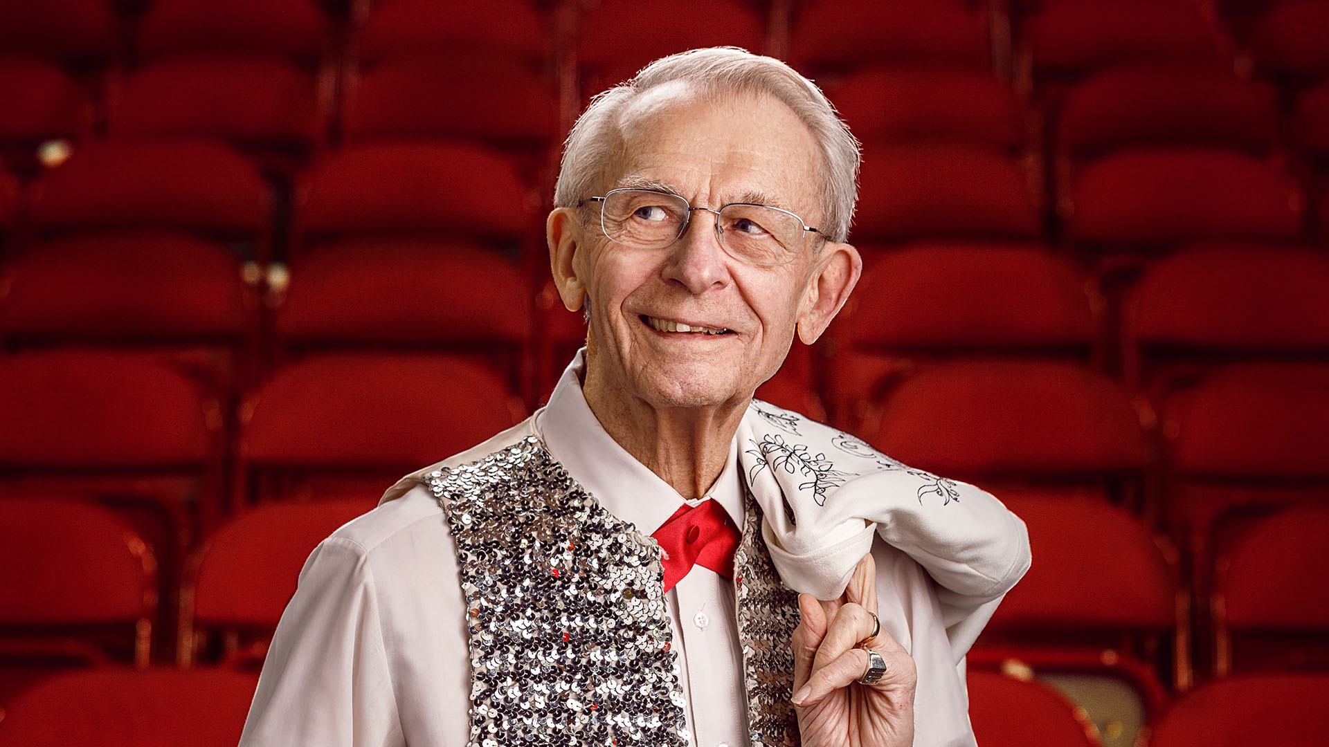 Headshot of Mike Leckrone. He is an older white man with short white hair, glasses, and silver eyes. He is smiling and looking to his right. He is wearing a white shirt, red tie, and a silver sequined vest