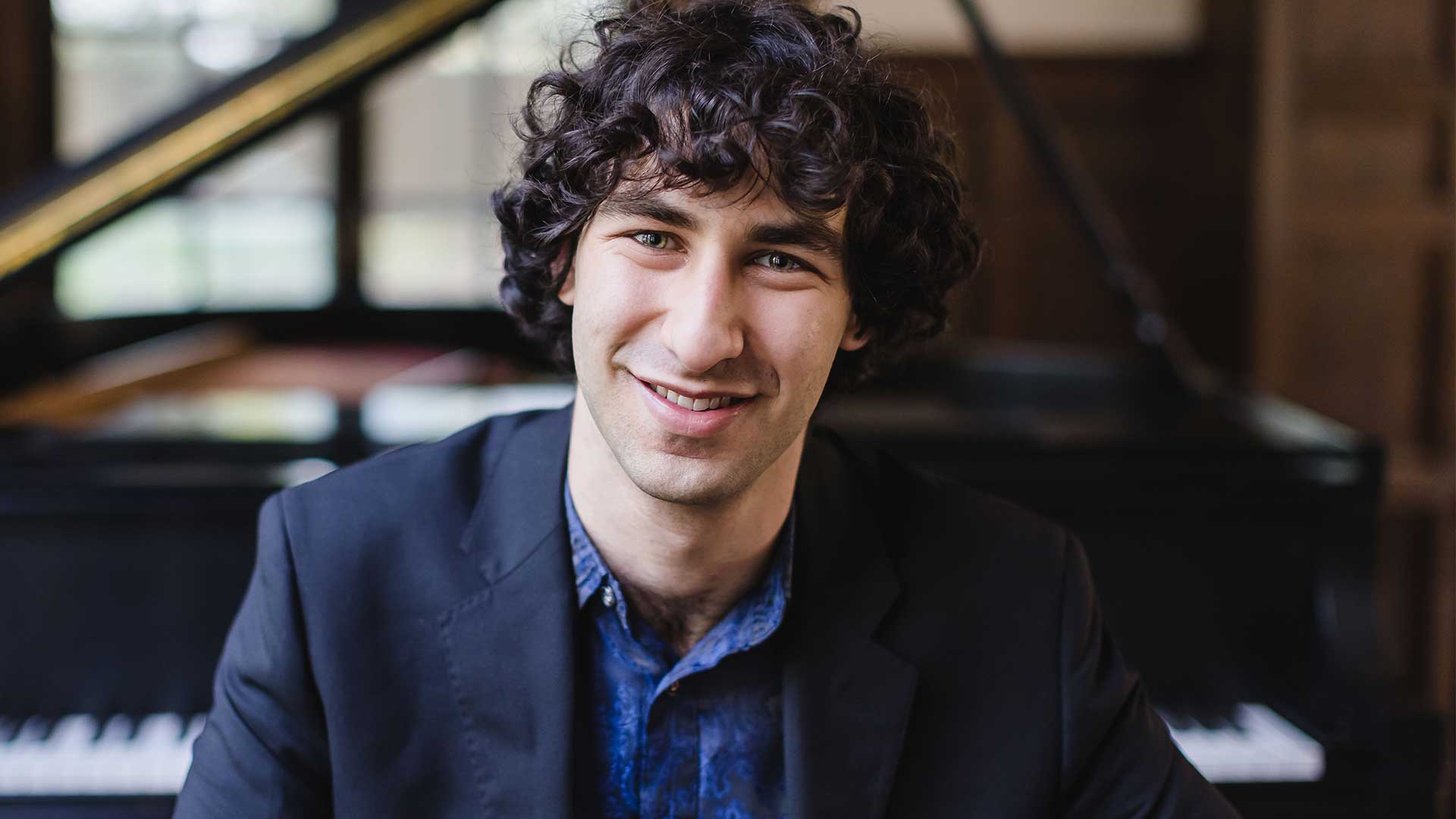 Headshot of a young caucasian man with medium length curly dark hair and greyish eyes. He is smiling and has a dark jacket on over a blue shirt.
