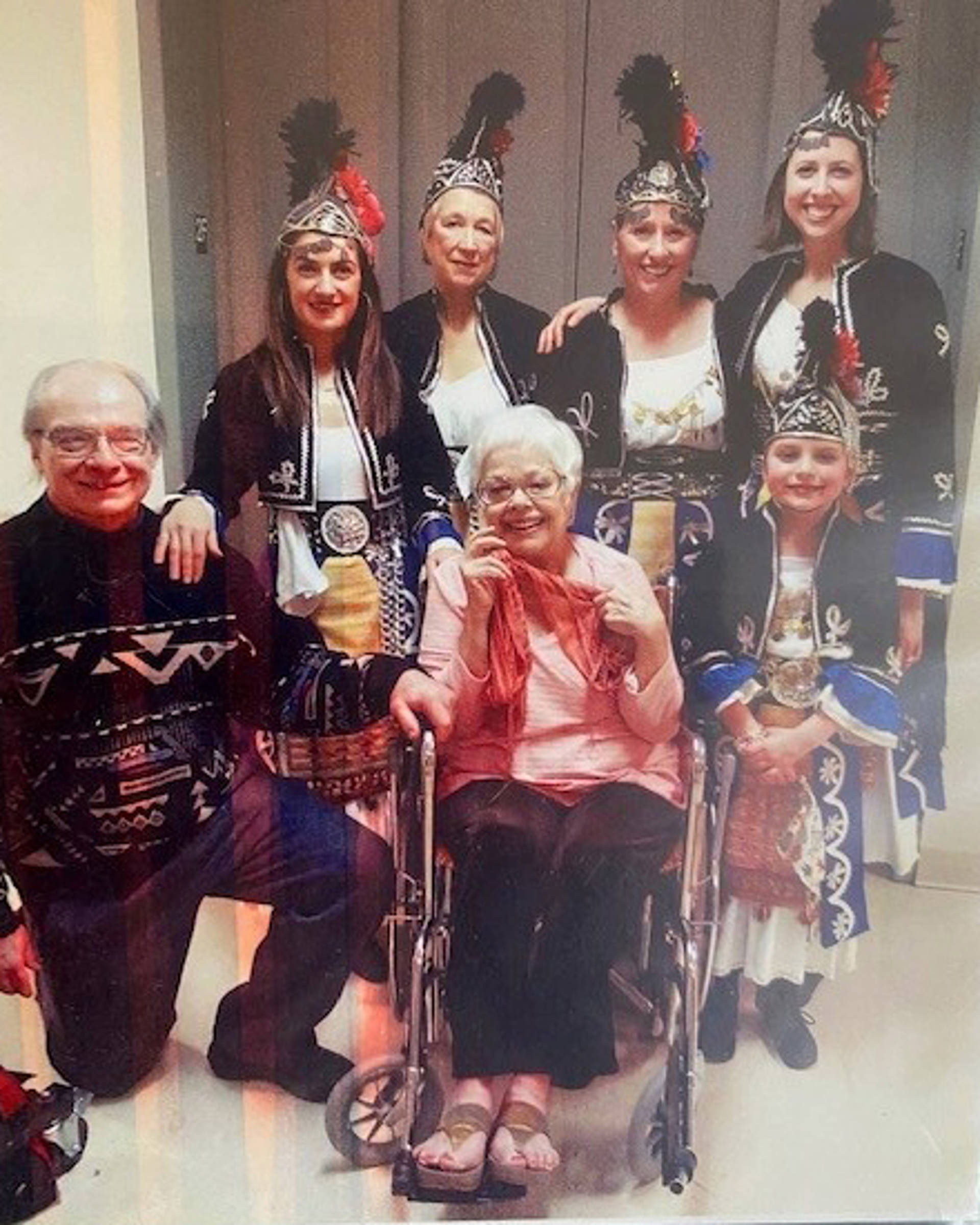 A group of people in traditional Greek dance outfits - black jackets and hats with silver ornaments and red highlights - standing behind an older woman with white hair sitting in a wheelchair.