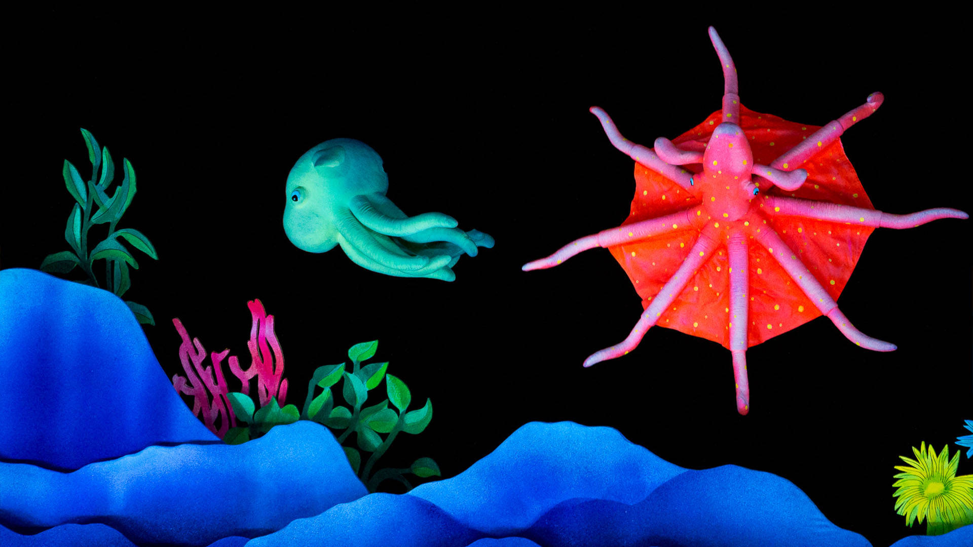 Puppets of a blue squid and red octopus swimming. Below them are blue rocks, green seaweed and pink coral.