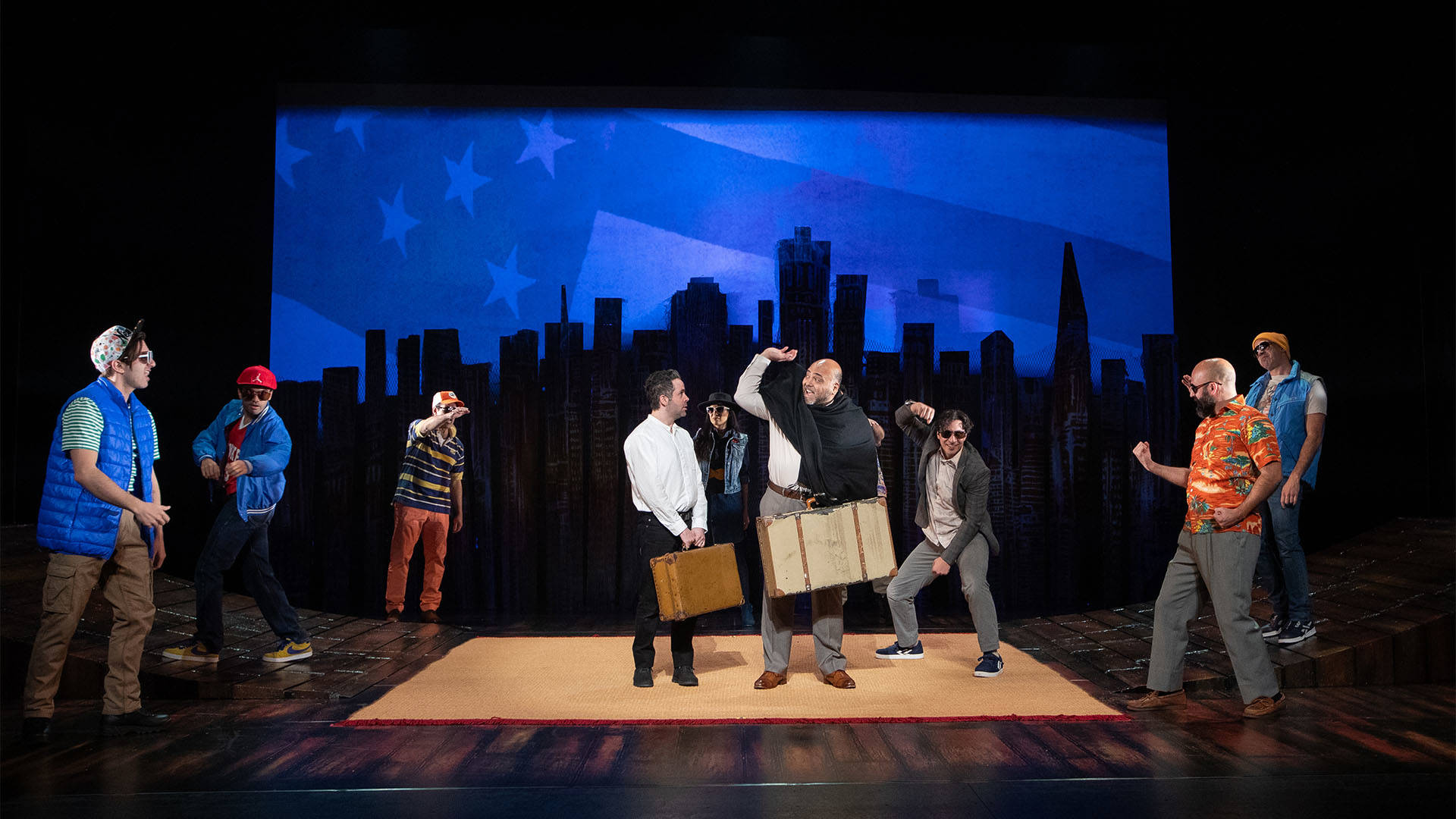 The cast of The Kite Runner in a variety of outfits on stage in front of a city skyline backdrop. Two in the center are holding brown leather suitcases while others move around them.