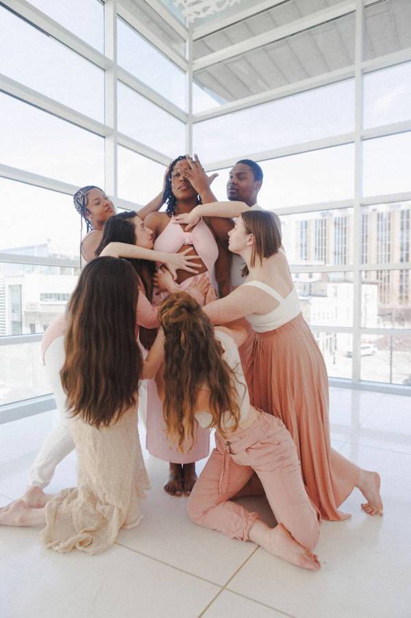 An african american woman  with dark hair and dark eyes in a light pink dress stands upright as 6 other dancers surround her and place their hands on her artistically. The background is large windows looking out on buildings.