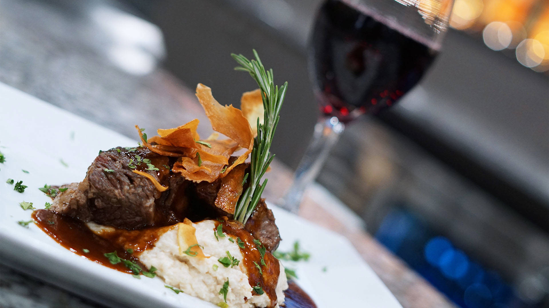 Close up image of braised short ribs on a bed of mashed potatoes with a blurry glass of red wine in the background.