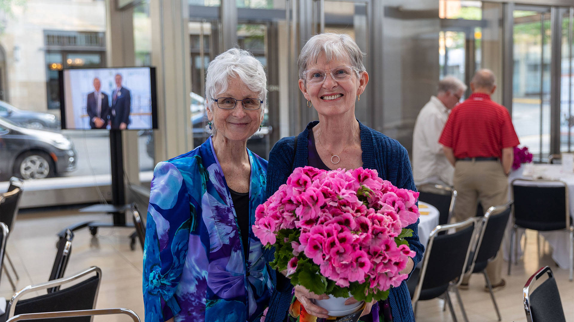 Two older caucasian women with short hair smile for a photo together. The woman on the right is holding a bouqet of pink flowers.
