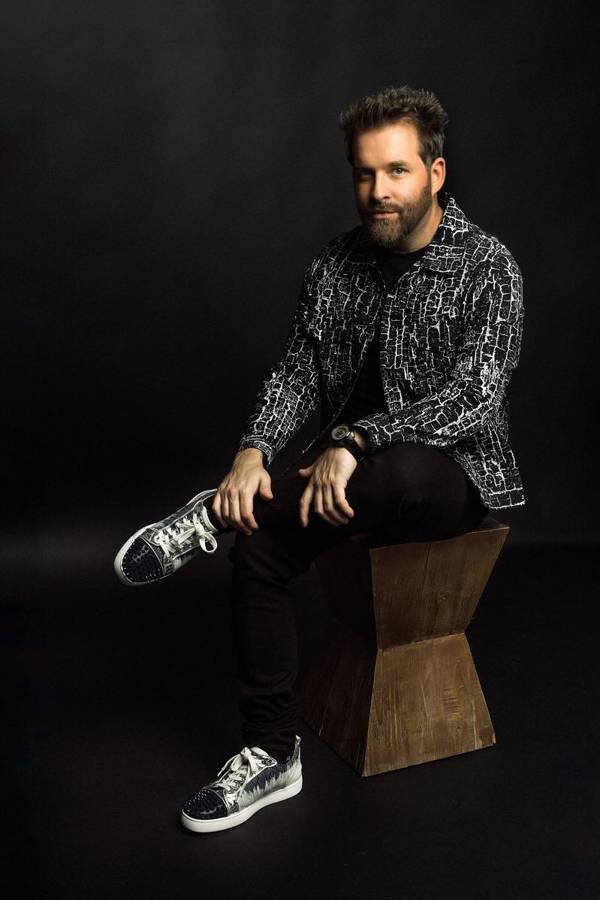 A young man with medium complexion, short brown hair, a beard, and a dark shirt. He is seated and crossing his left leg.