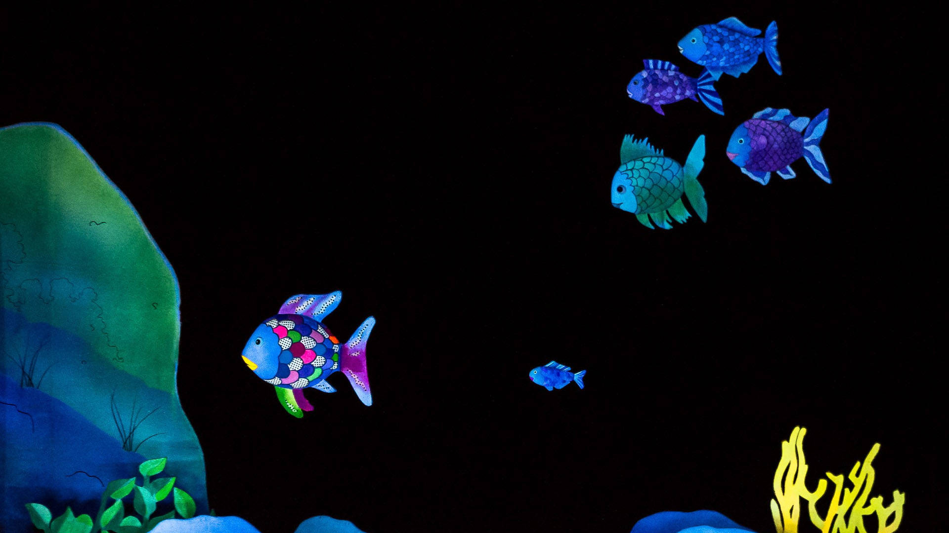 Puppets of fish. One has rainbow scales and is staring at a large rock. Five smaller fish of various shades of blue and purple follow the rainbow fish.