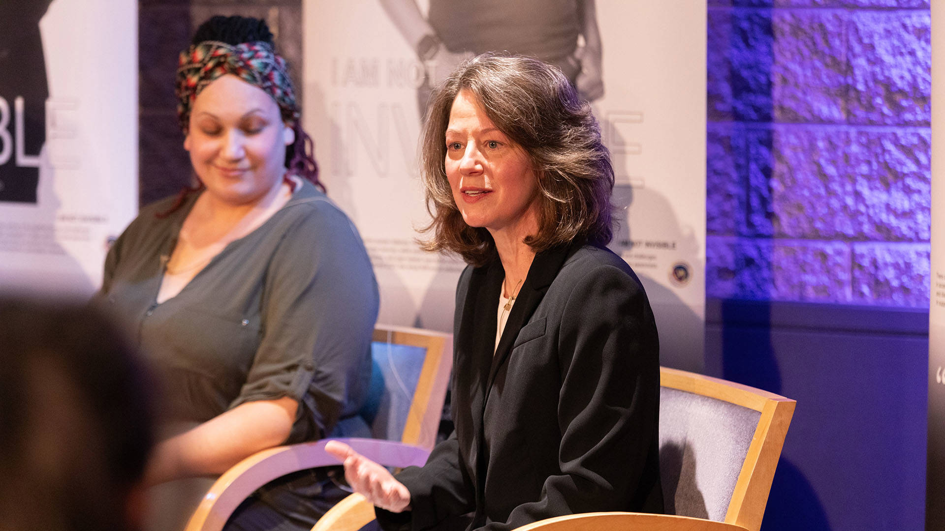 A woman with greying brown hair, light complexion and a dark top sitting and speaking.