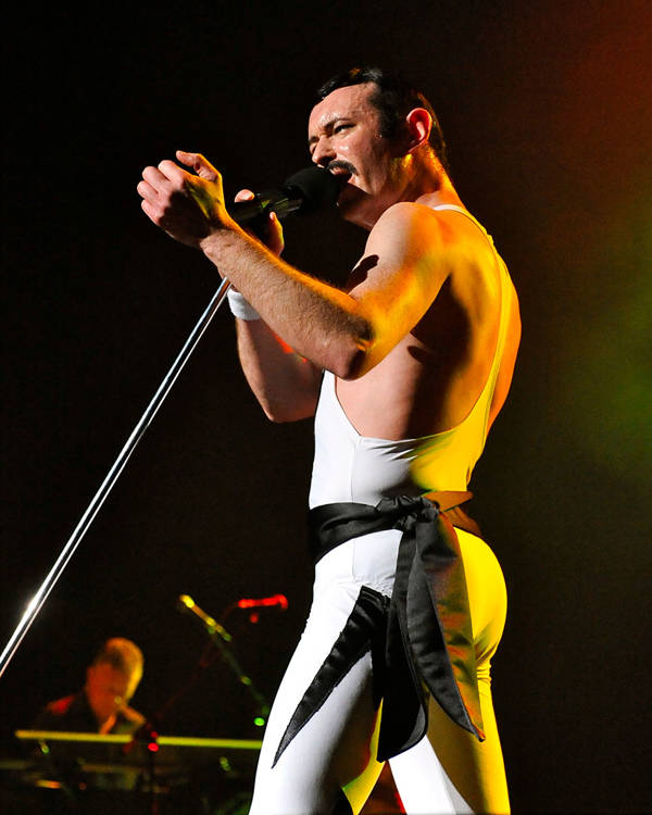 A caucasian man with short black hair and a moustache singing into a microphone with his right hand outstretched. He is wearing all white with a tank top.
