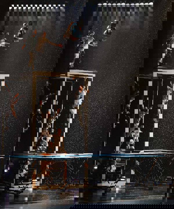 A group of dancers scaling a large framed box. One is doing a flip above a large trampoline on stage while the others watch in excitement.