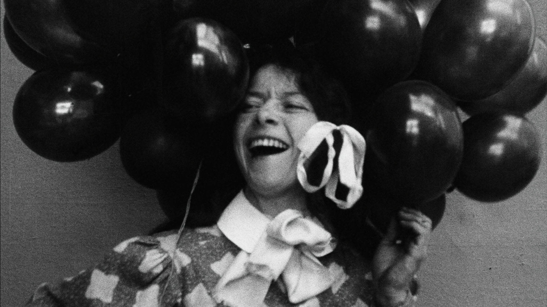 Vintage black and white headshot of a light complexion woman with dark hair. Her mouth is wide open in a large smile as she holds a bouquet of balloons above her head like a hat.
