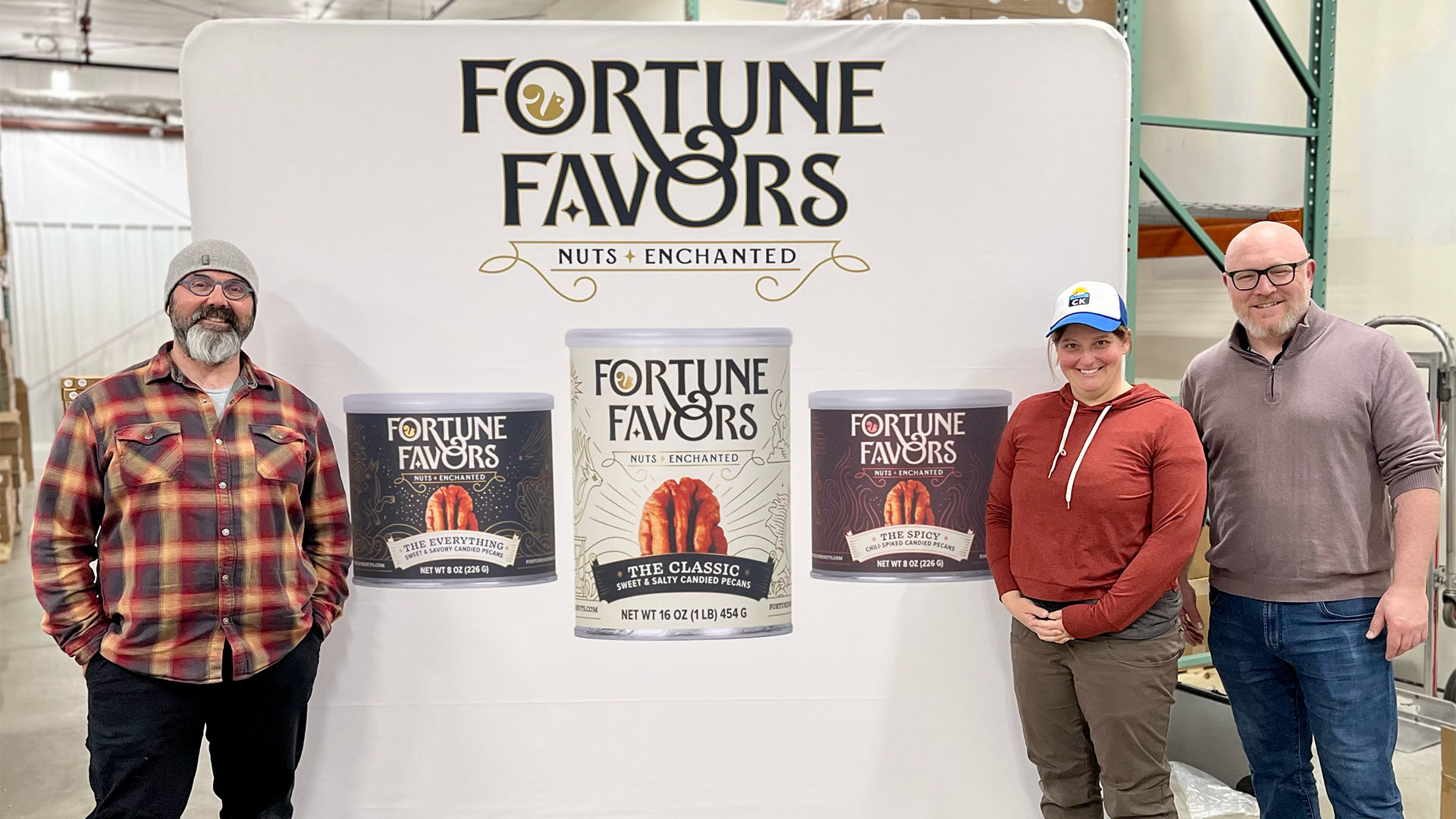 Two middle aged men and a younger woman pose for a photo in front of a large banner with images of cans of nuts and the brand name "Fortune Favors"